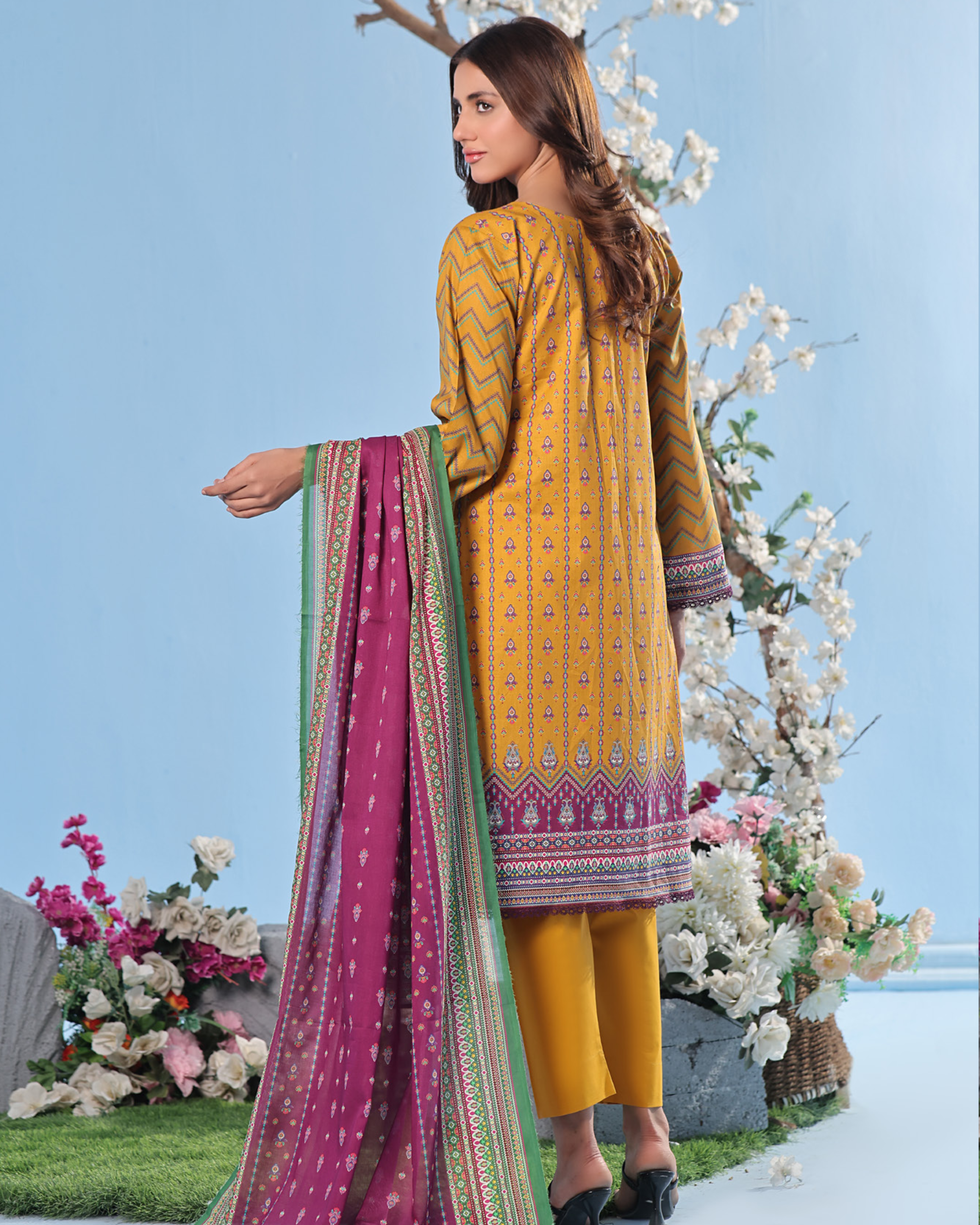 Woman in yellow traditional outfit with a pink dupatta, standing against a floral and blue sky background.