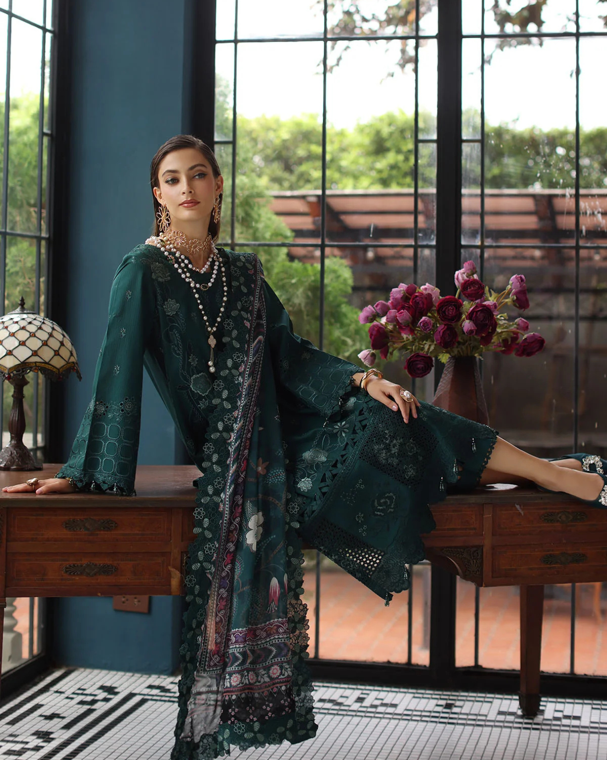 Woman in a dark green embroidered dress sitting on a wooden desk with a decorative background.