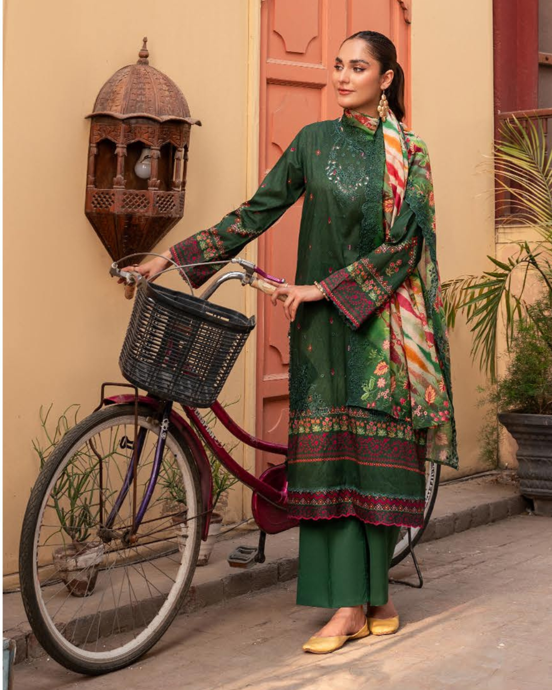 Woman in green traditional outfit with a colorful scarf, standing next to a bicycle in an outdoor setting.
