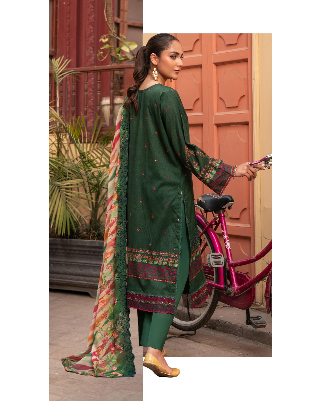 Woman in green traditional outfit with a colorful scarf, standing next to a bicycle in an outdoor setting.