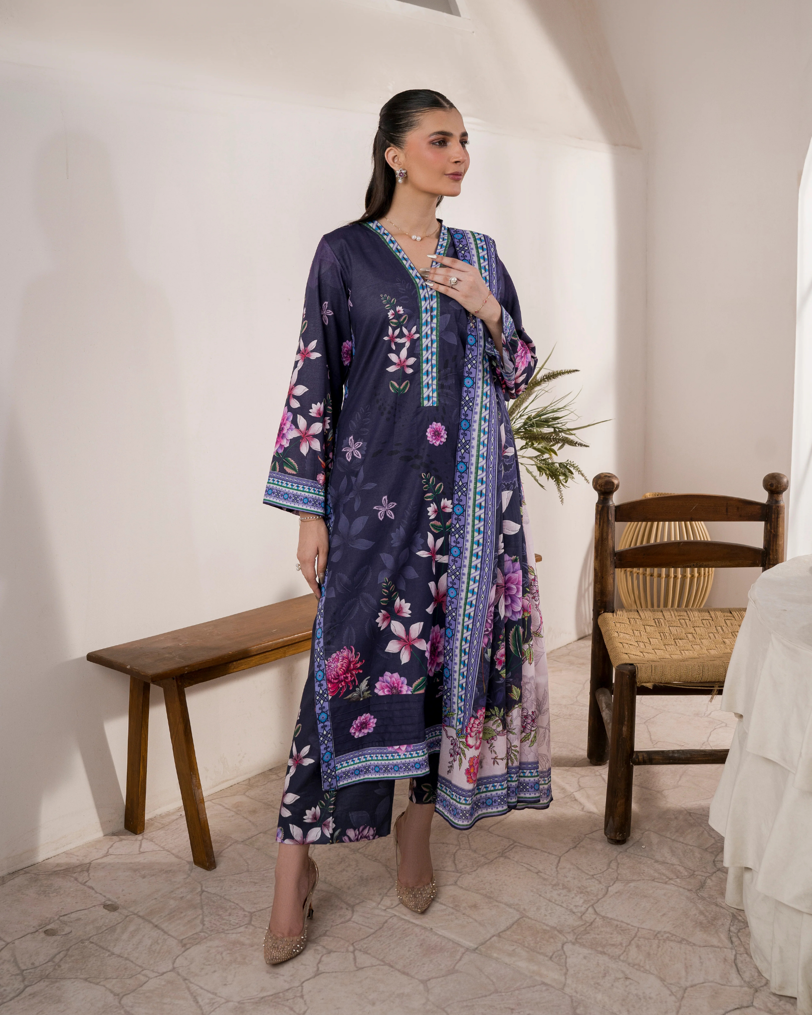 Woman wearing a long, dark blue dress with floral patterns in a room with white walls and wooden furniture.