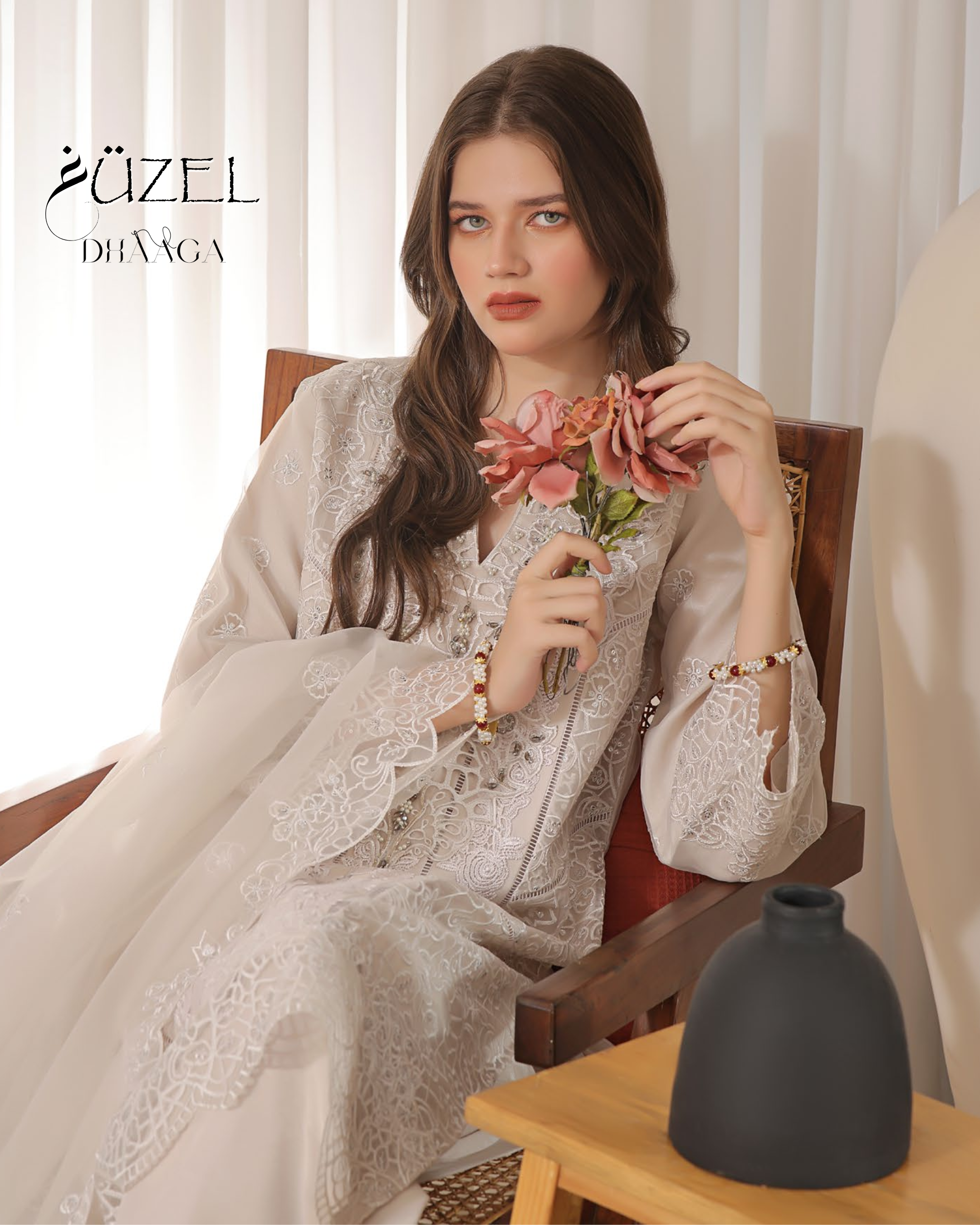 Woman in a white embroidered outfit holding flowers, sitting on a chair with a vase on a table.