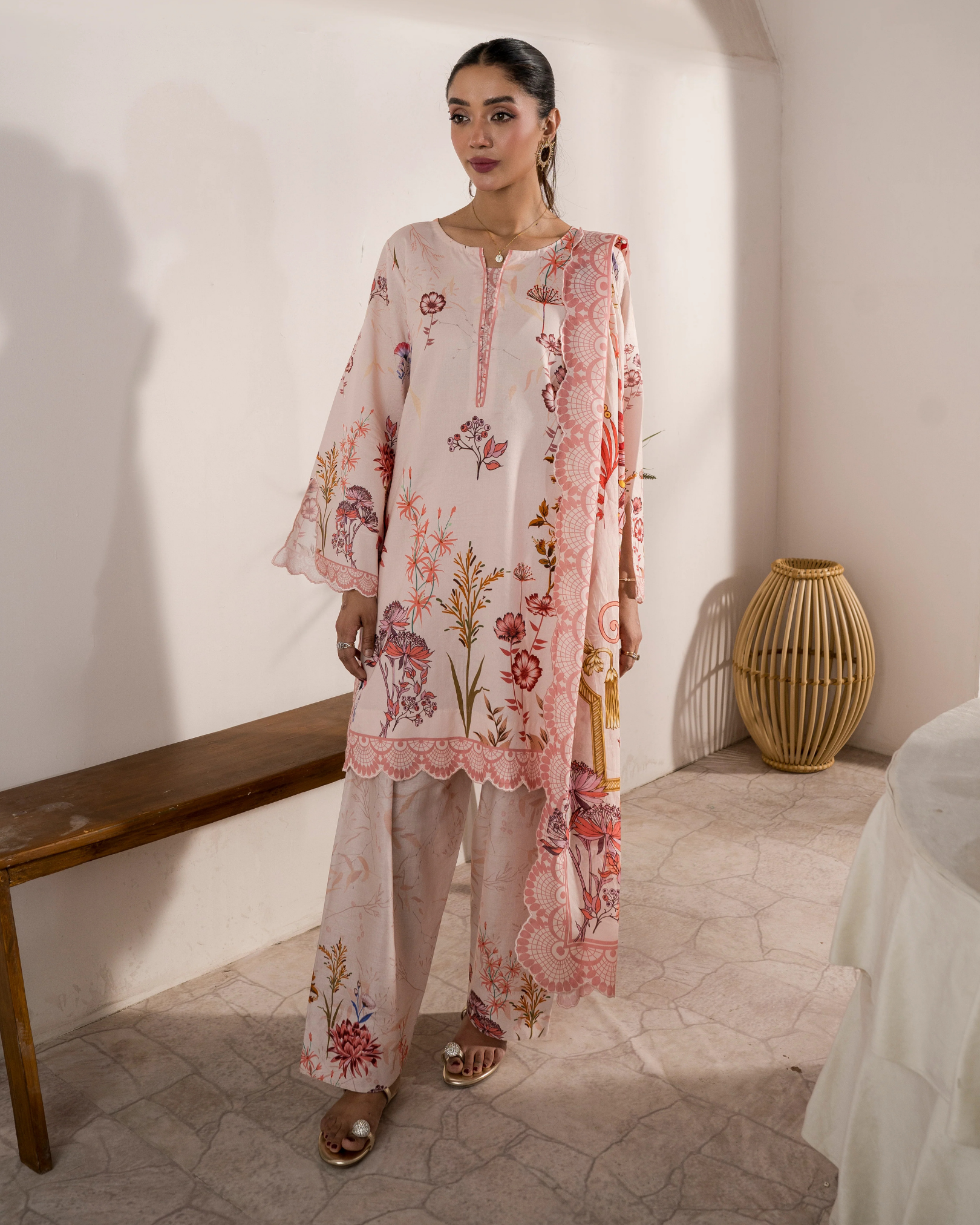 Woman wearing a floral outfit standing in a room with a wooden bench and woven basket.