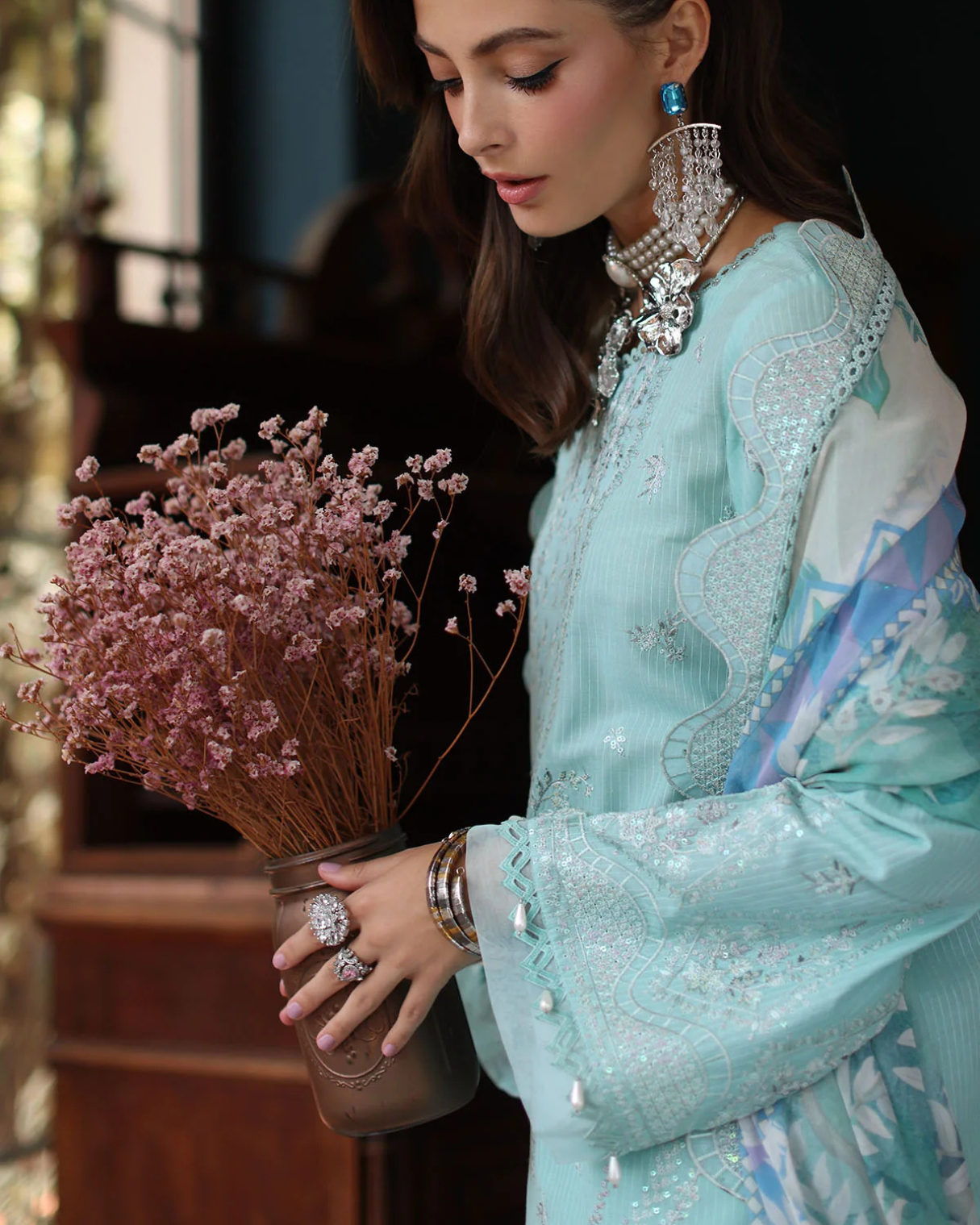 Woman in a light blue embroidered outfit holding pink flowers indoors.