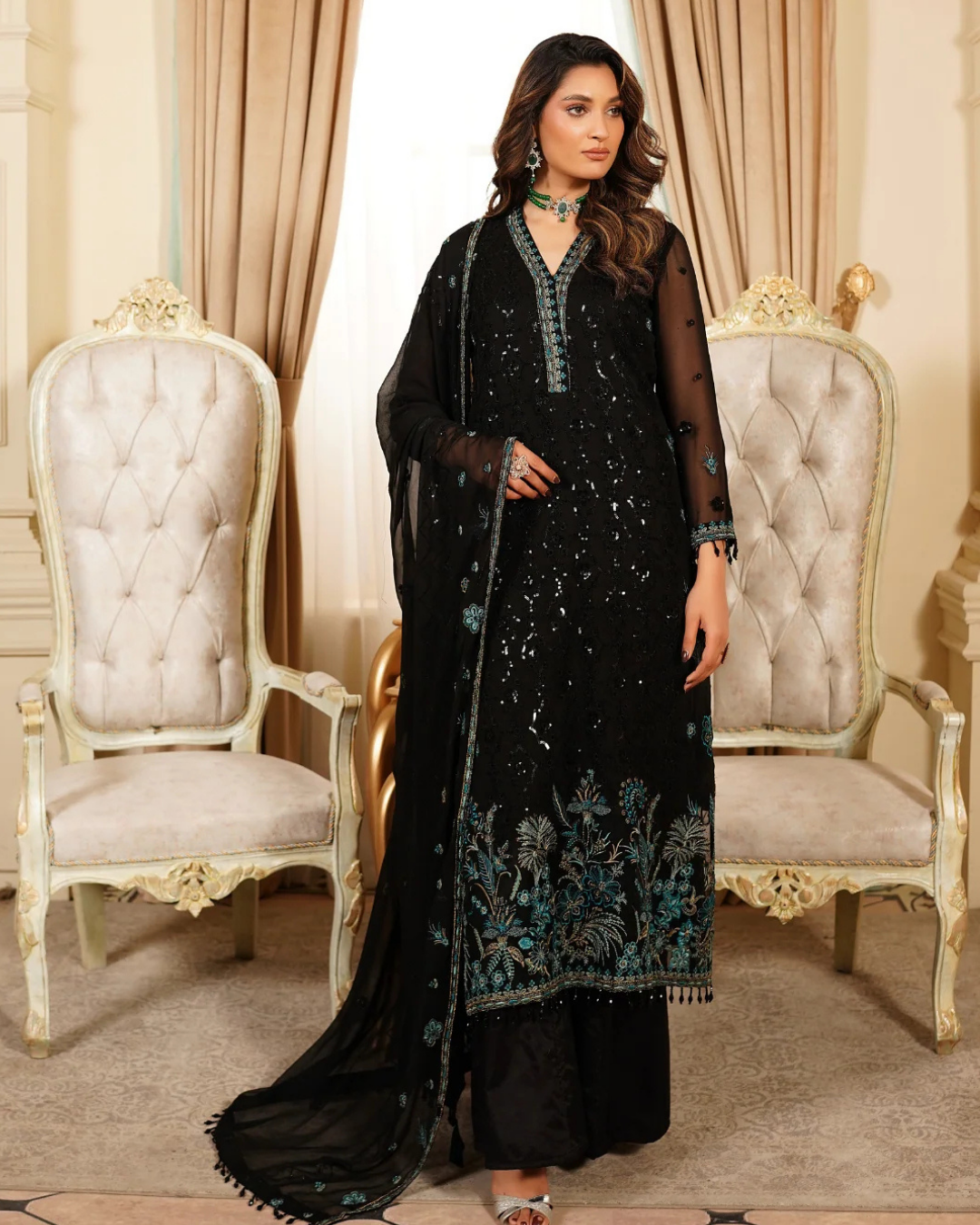 Woman in a black embroidered outfit standing in an elegant room with ornate chairs and curtains.