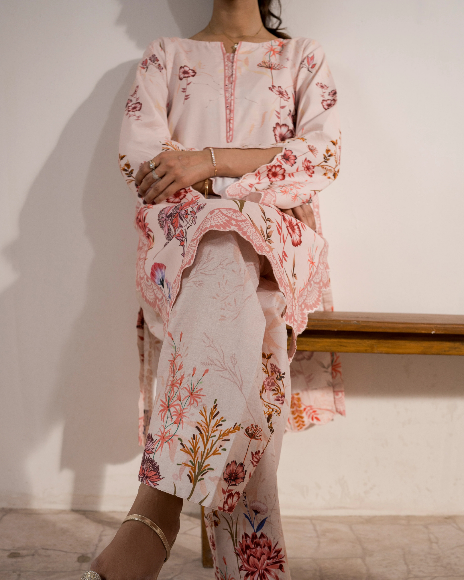 Person wearing a floral outfit sitting on a bench against a white wall.