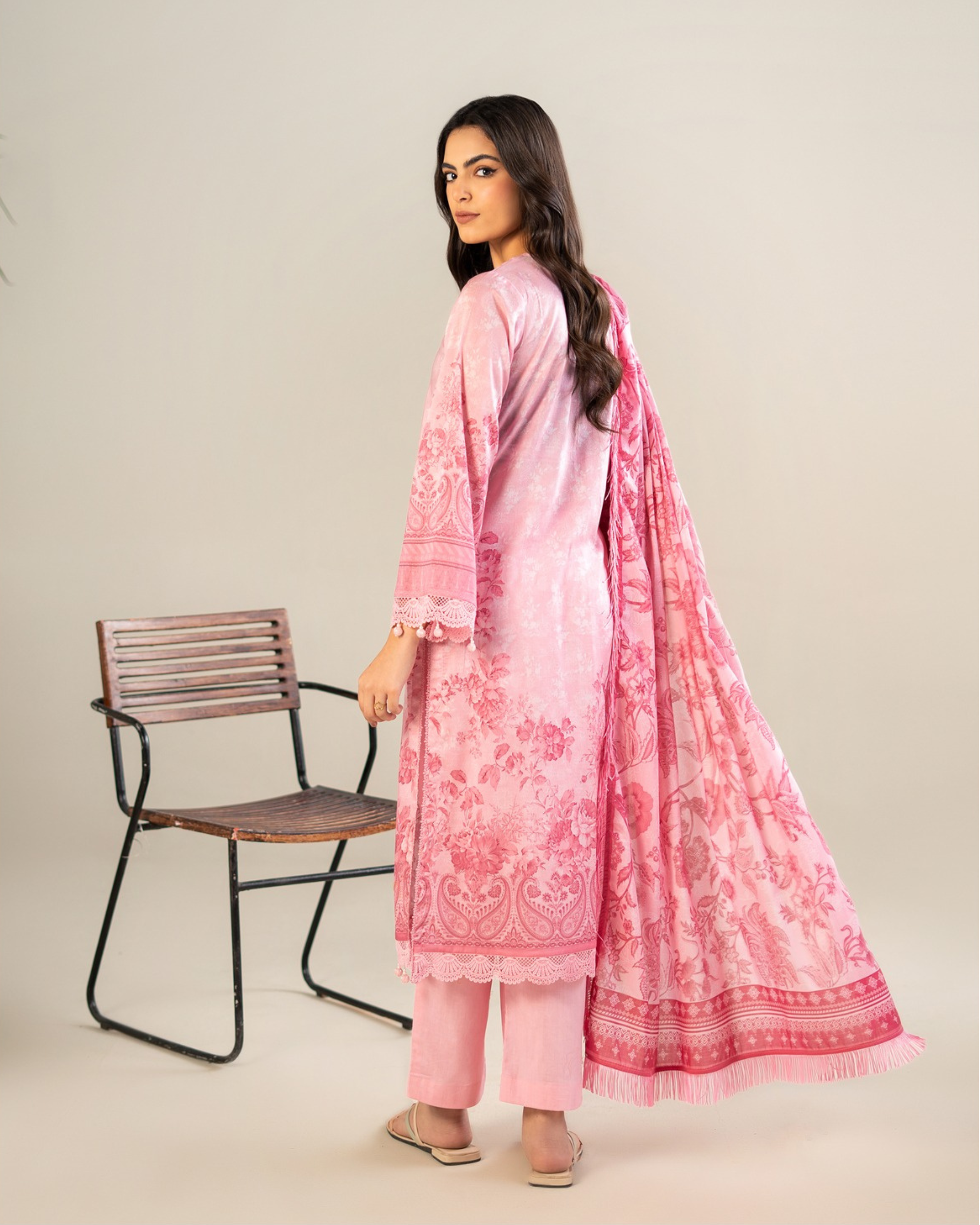Woman wearing a pink embroidered outfit with a matching dupatta, standing next to a chair on a plain background.