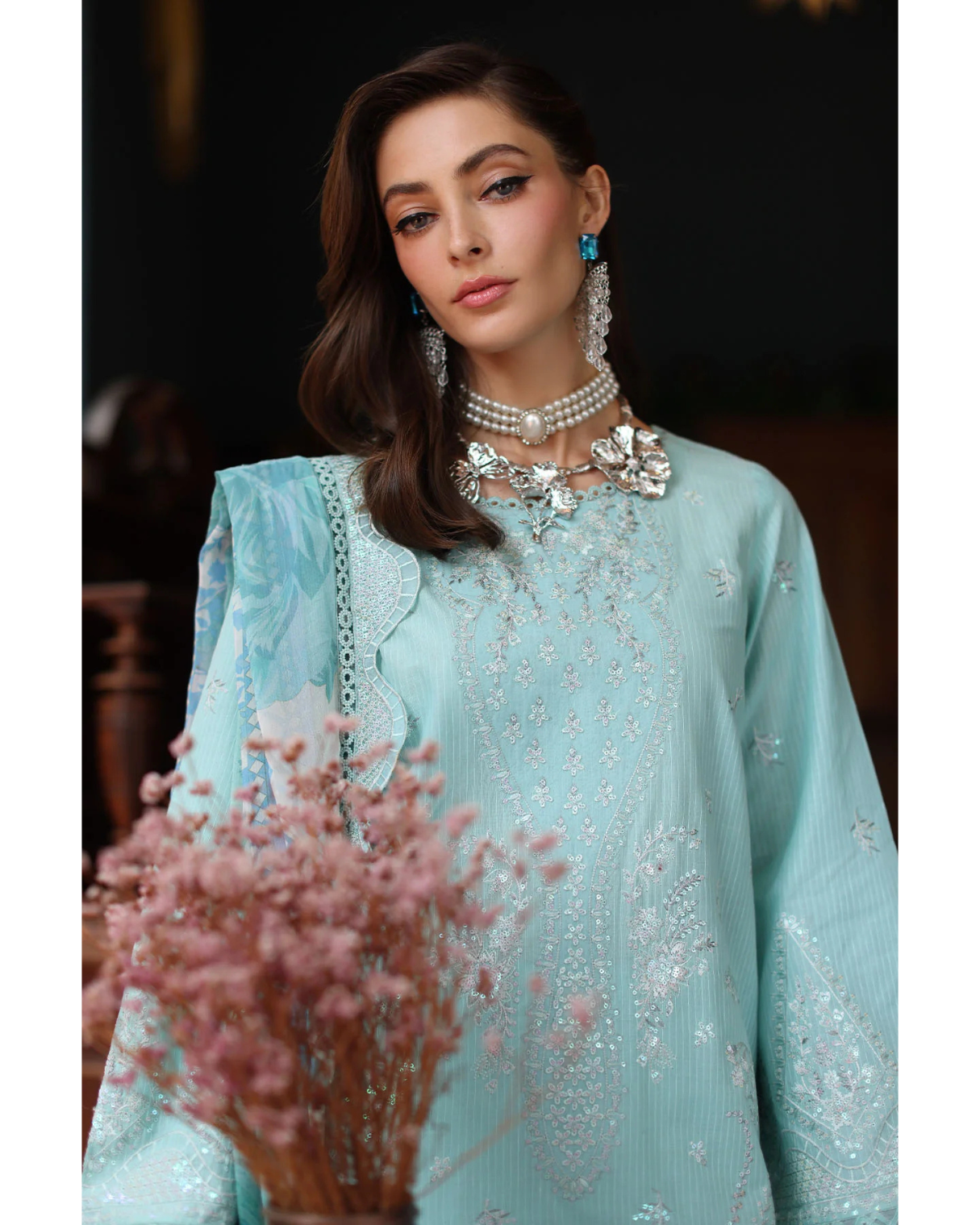 Woman wearing a light blue embroidered dress with jewelry, standing next to pink flowers.