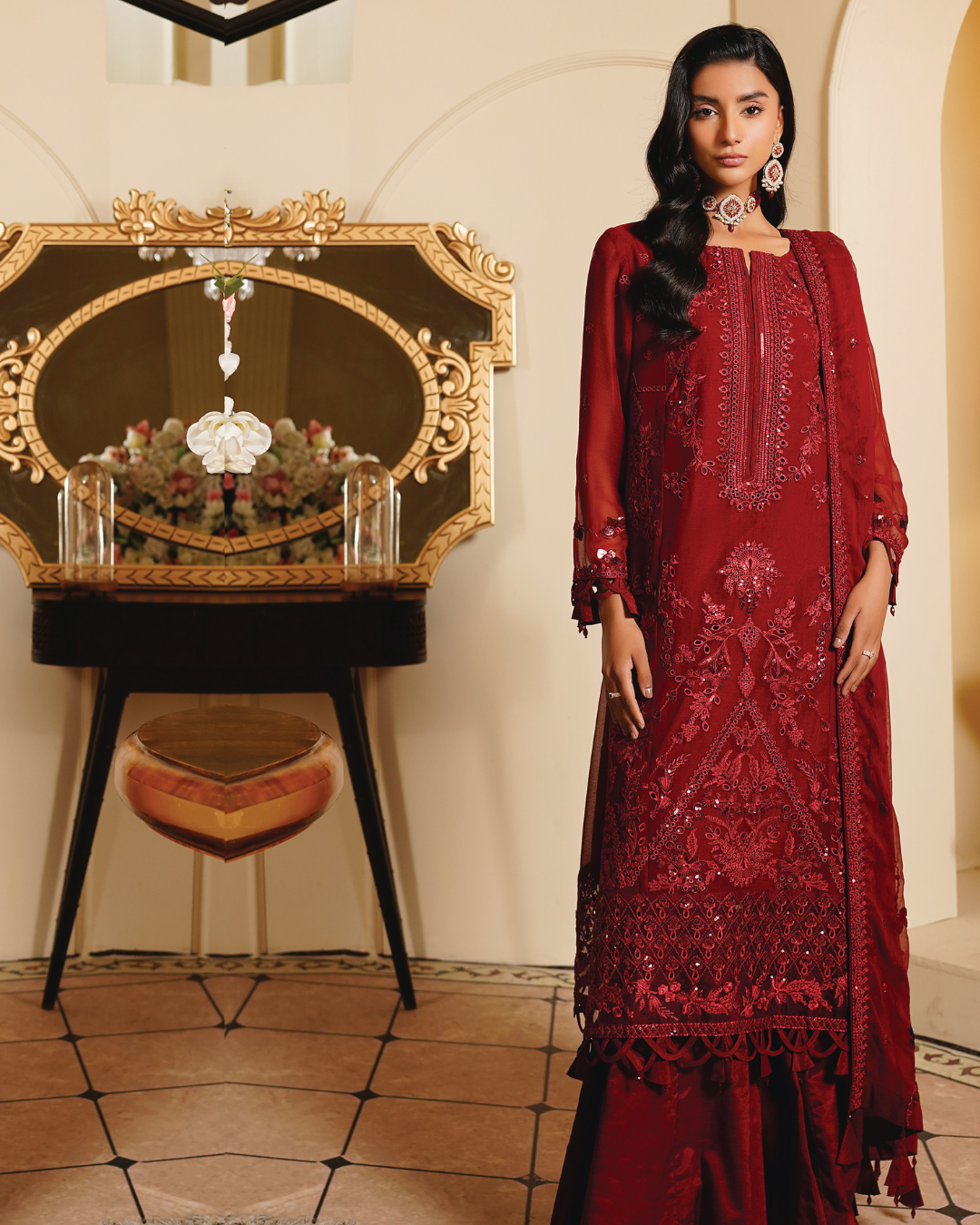 Woman in a red embroidered outfit standing in a decorated room with a mirror and table.