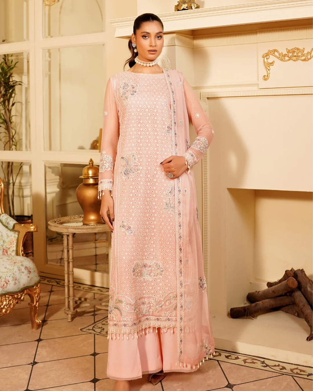 Woman in a pink embroidered outfit standing in an elegant room with decorative elements.