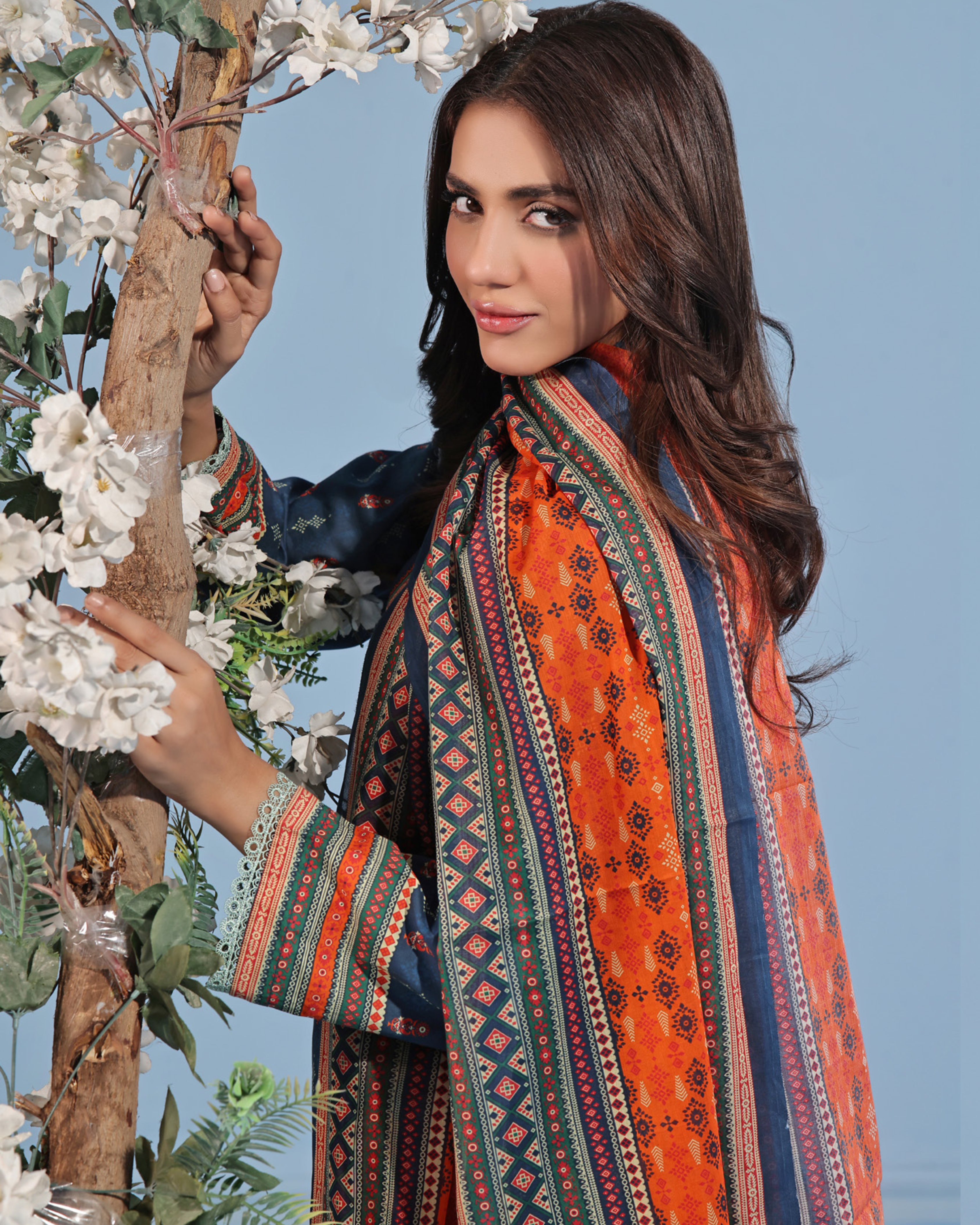 Woman wearing a colorful traditional outfit with a patterned shawl, holding flowers against a blue sky.