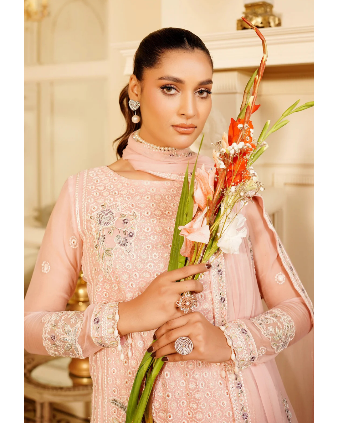 Woman in a pink embroidered outfit holding flowers indoors.