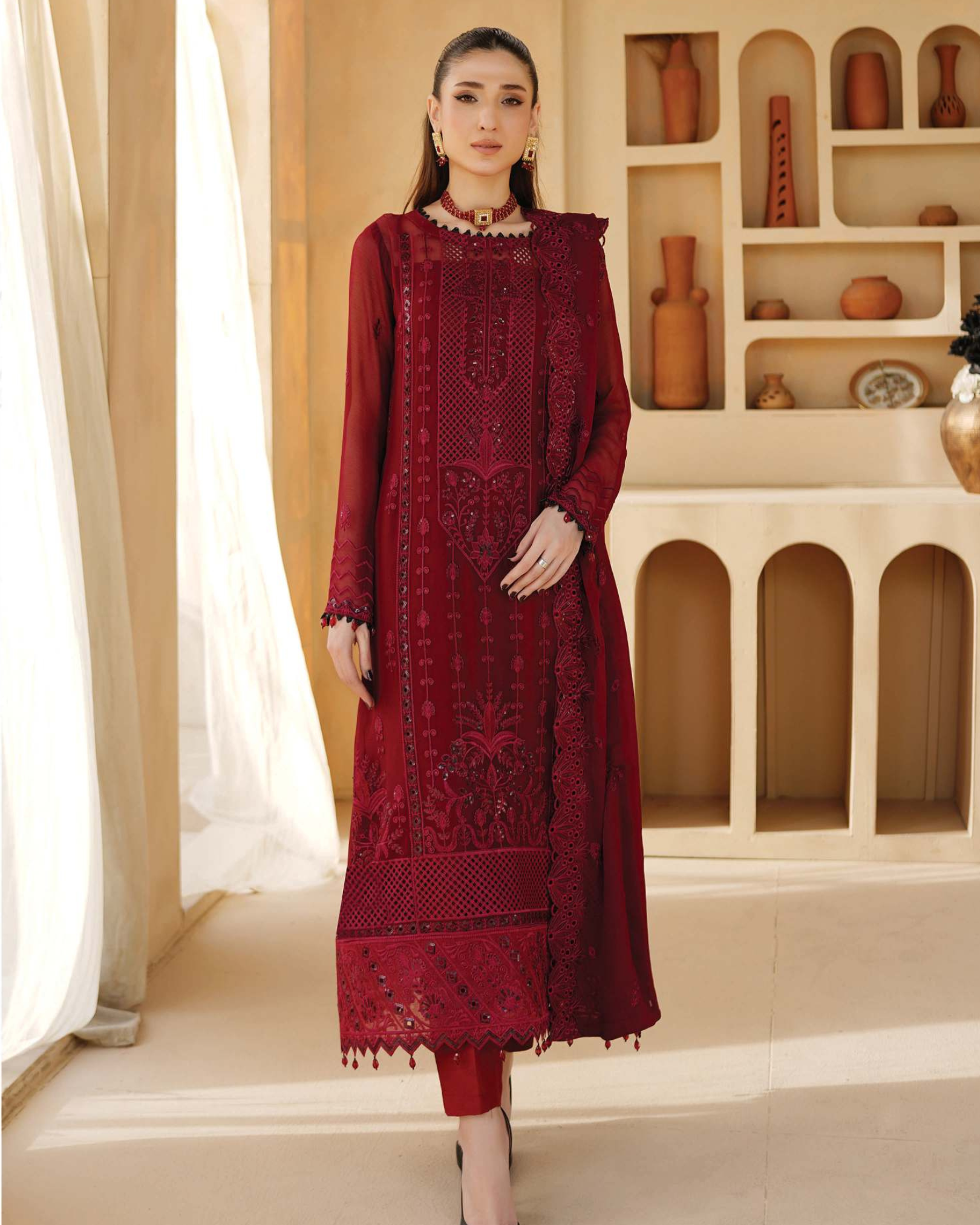 Woman wearing a red embroidered outfit standing in a room with decorative shelves.