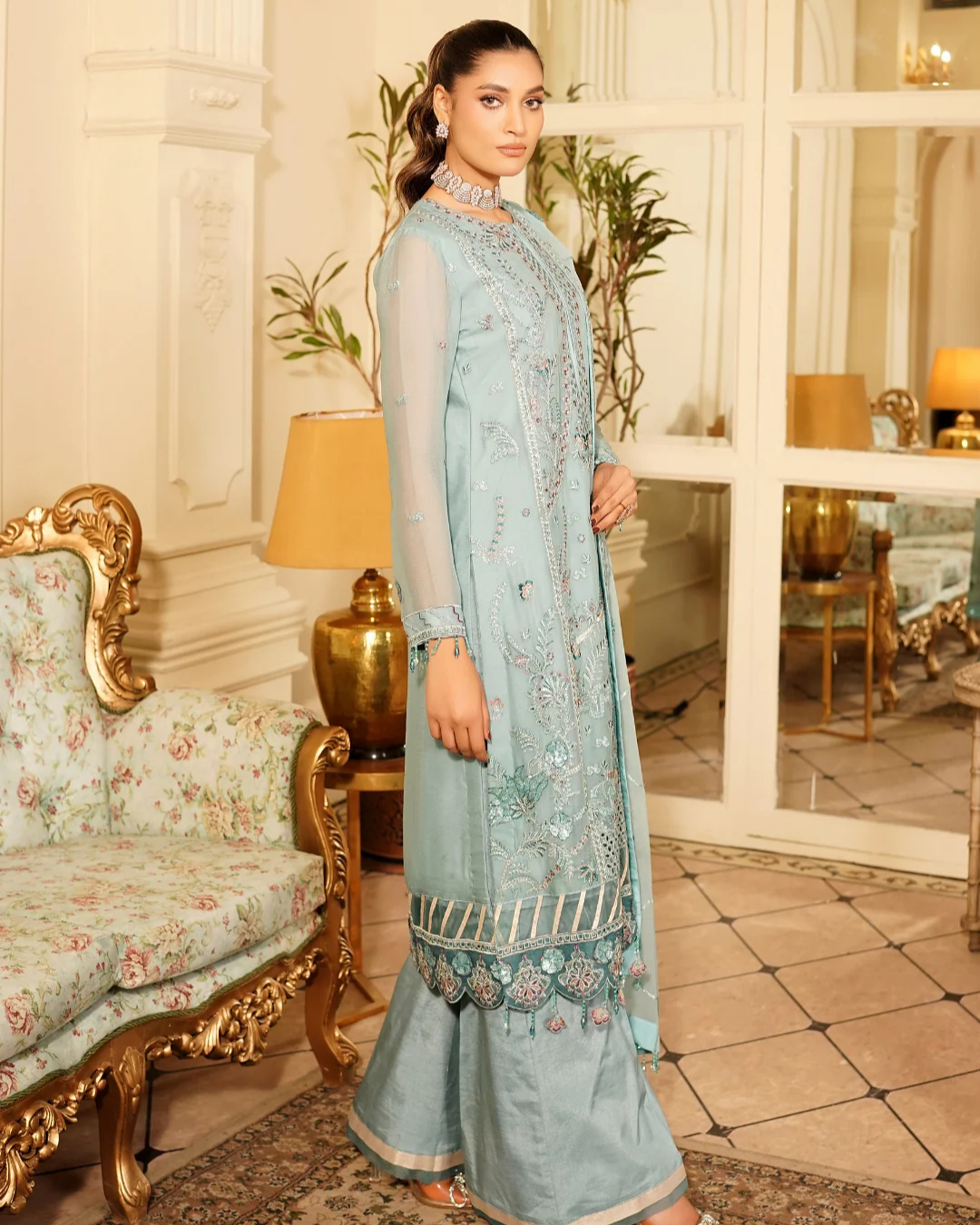 Woman in a light blue traditional outfit standing in an elegant room with decorative furniture.