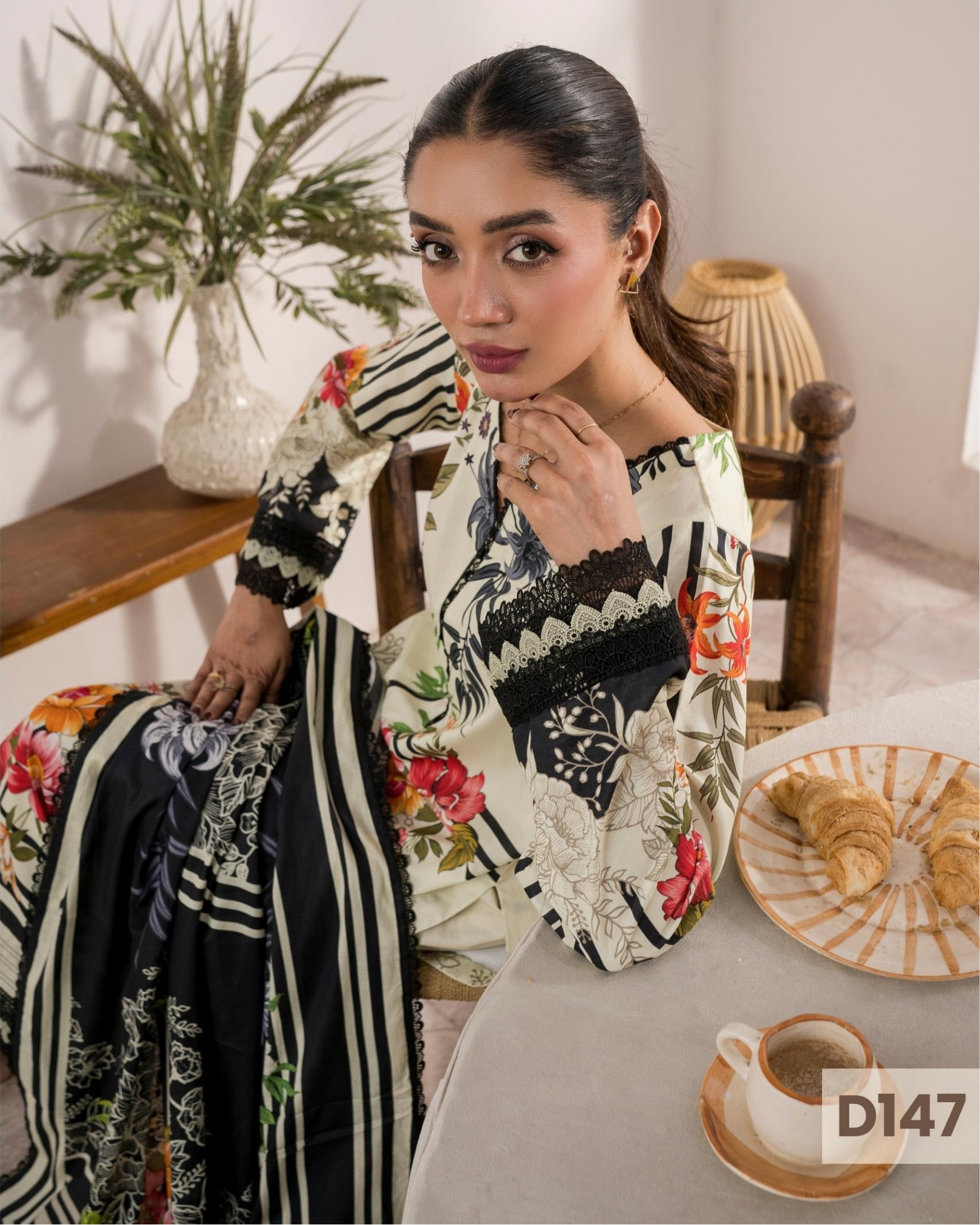 Woman sitting at a table with a floral and geometric patterned dress, surrounded by a cozy indoor setting.