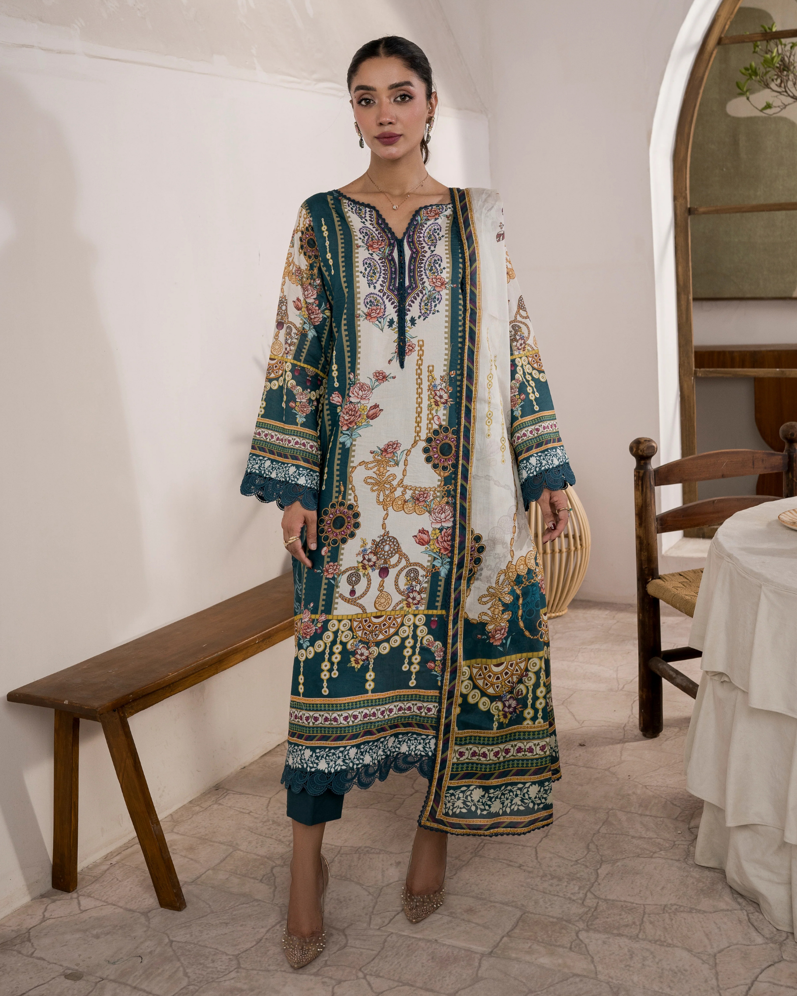 Woman wearing a patterned dress in an indoor setting with wooden furniture and a white wall.