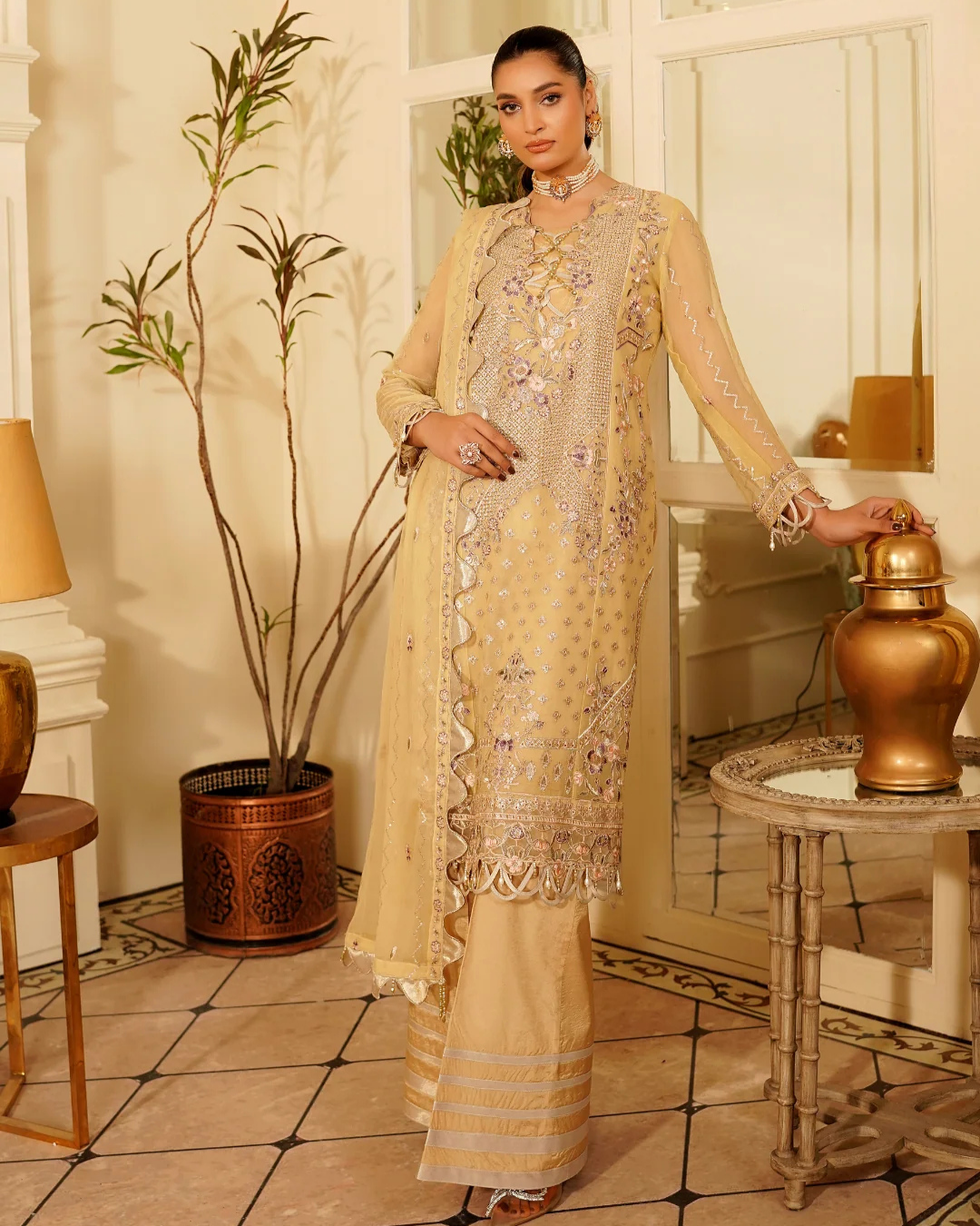Woman in a beige embroidered outfit standing in an elegant room with decorative elements.