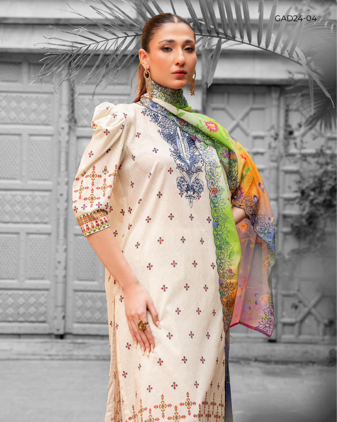 Woman wearing a cream-colored embroidered outfit with a colorful scarf against a decorative wall.