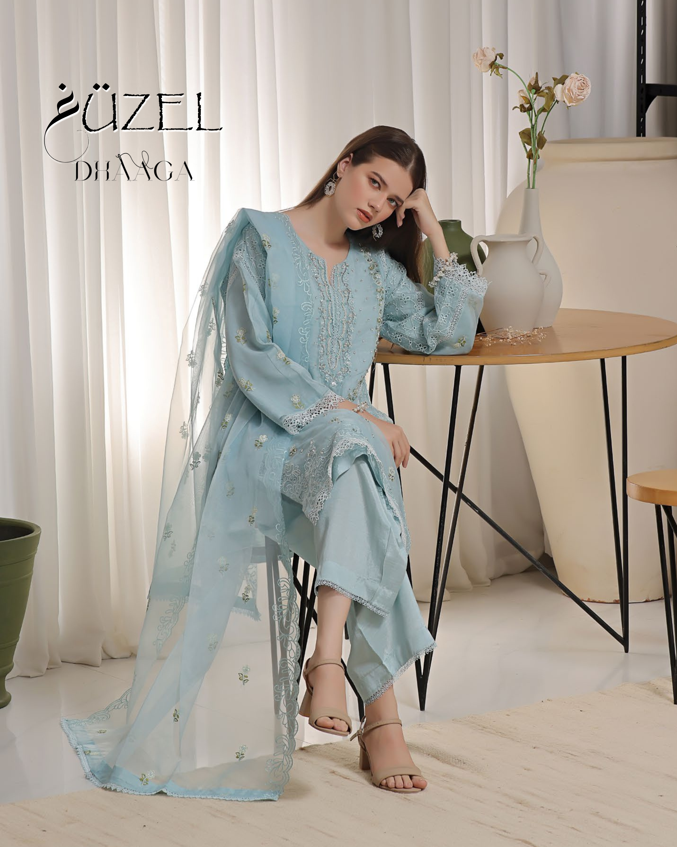 Woman in a light blue embroidered outfit sitting in a room with white curtains and a table.