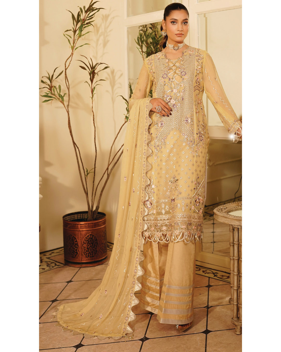 Woman in a beige embroidered traditional outfit standing in a room with decorative elements.