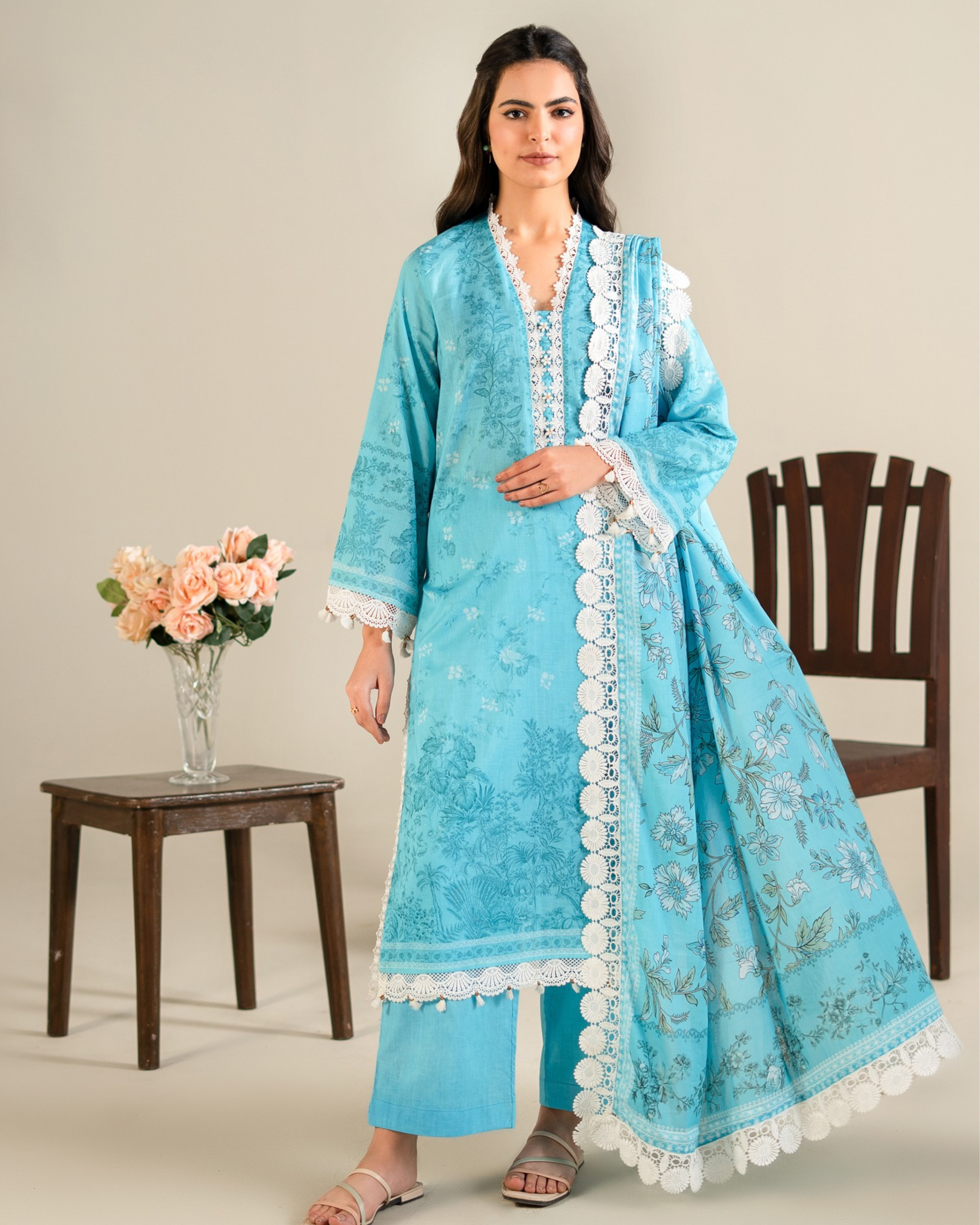 Woman wearing a blue embroidered traditional outfit with white lace details, standing next to a wooden chair and table with flowers.