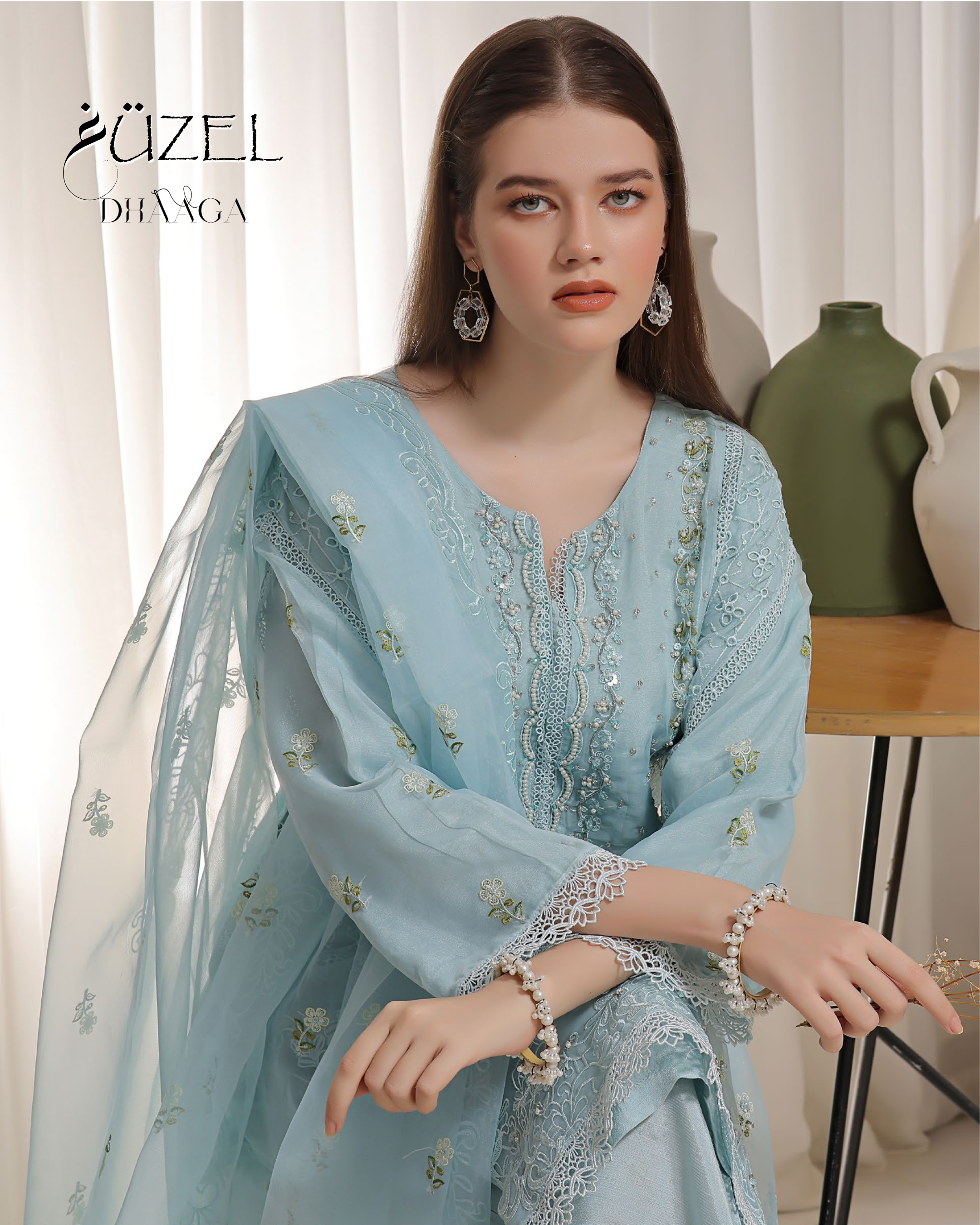 Woman wearing a light blue embroidered dress with a white dupatta, sitting indoors.
