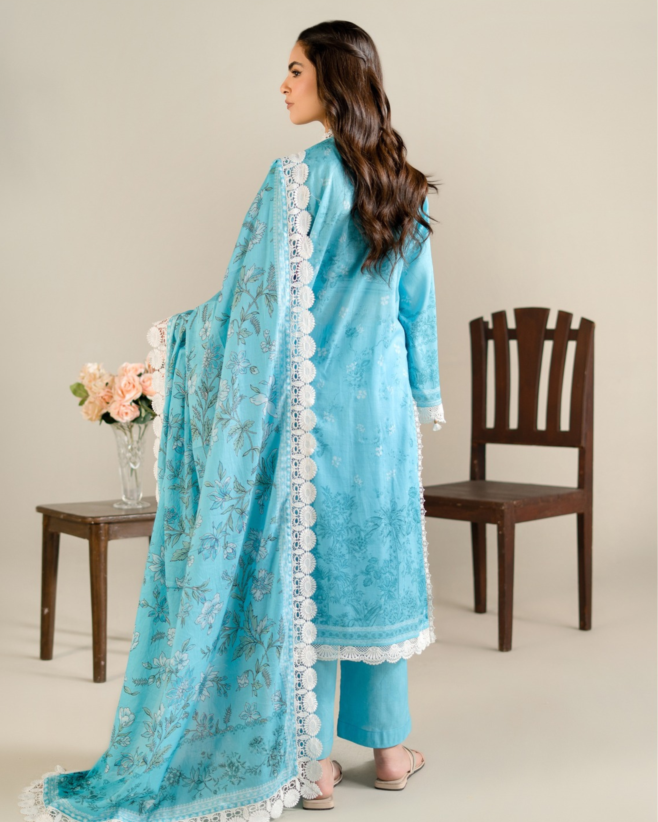 Woman wearing a blue traditional outfit with white lace details, standing next to a wooden chair and table with flowers.