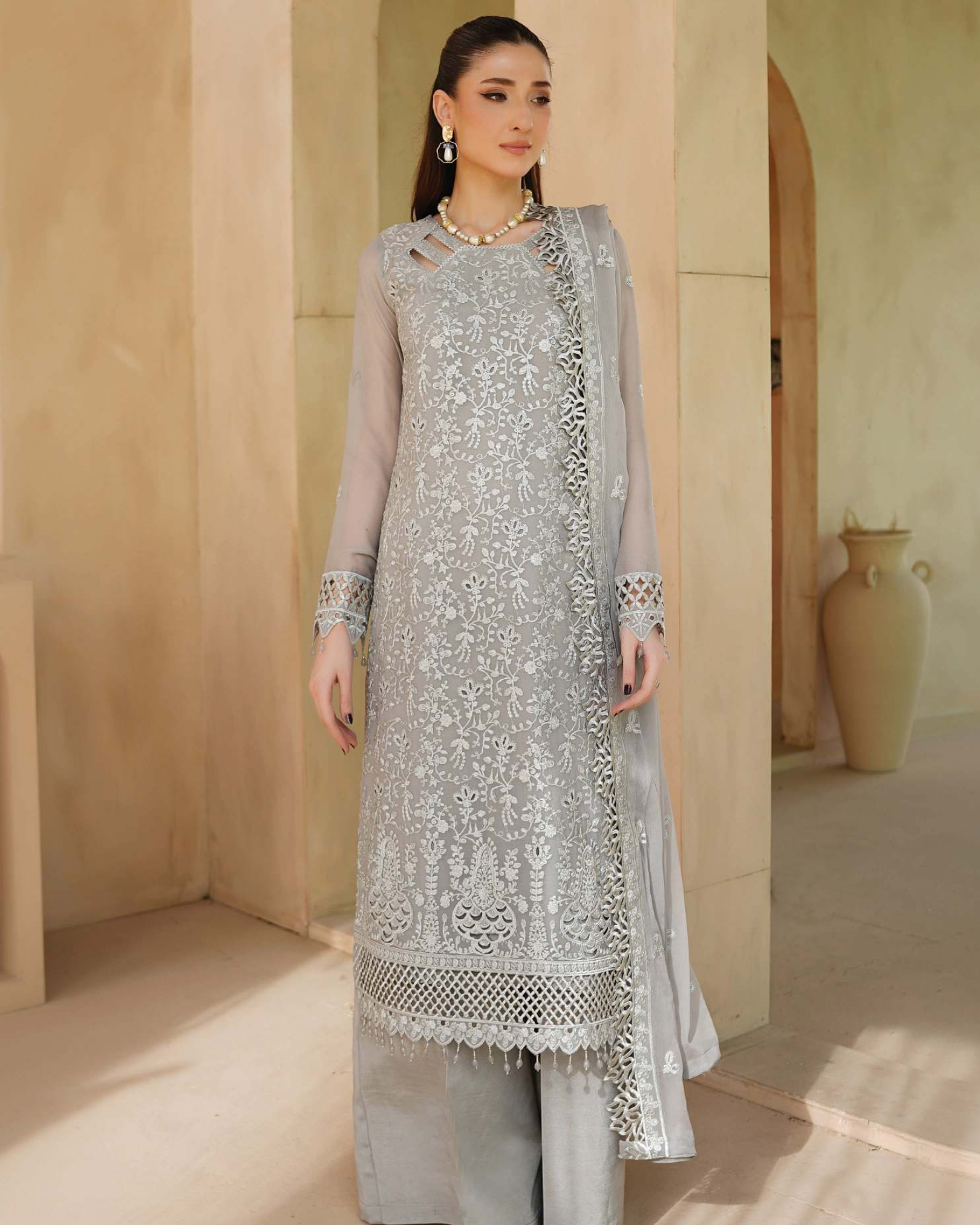 Woman wearing a light gray embroidered traditional outfit in a warm-toned room.