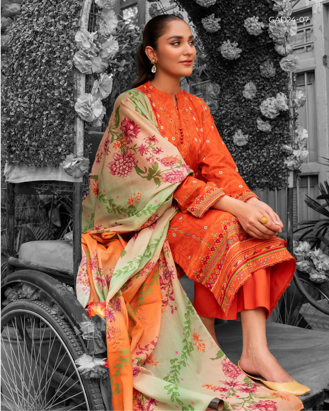 Woman in traditional orange and floral outfit sitting outdoors with a black and white background
