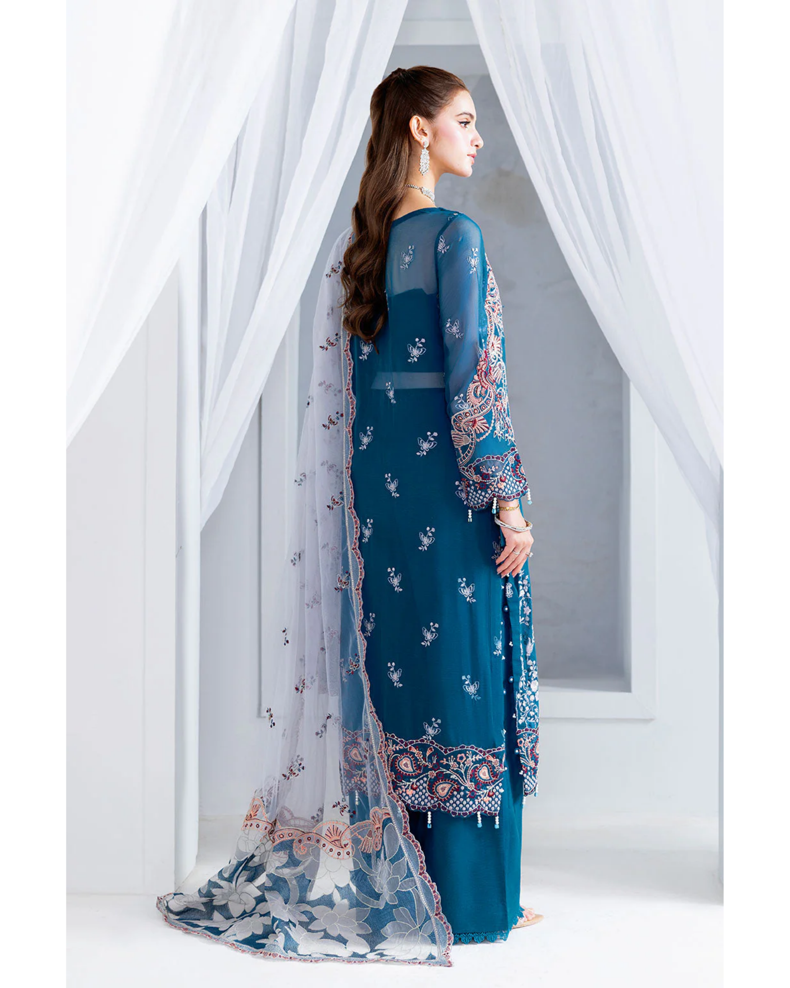 Woman wearing a blue embroidered traditional outfit with a sheer overlay against white curtains.