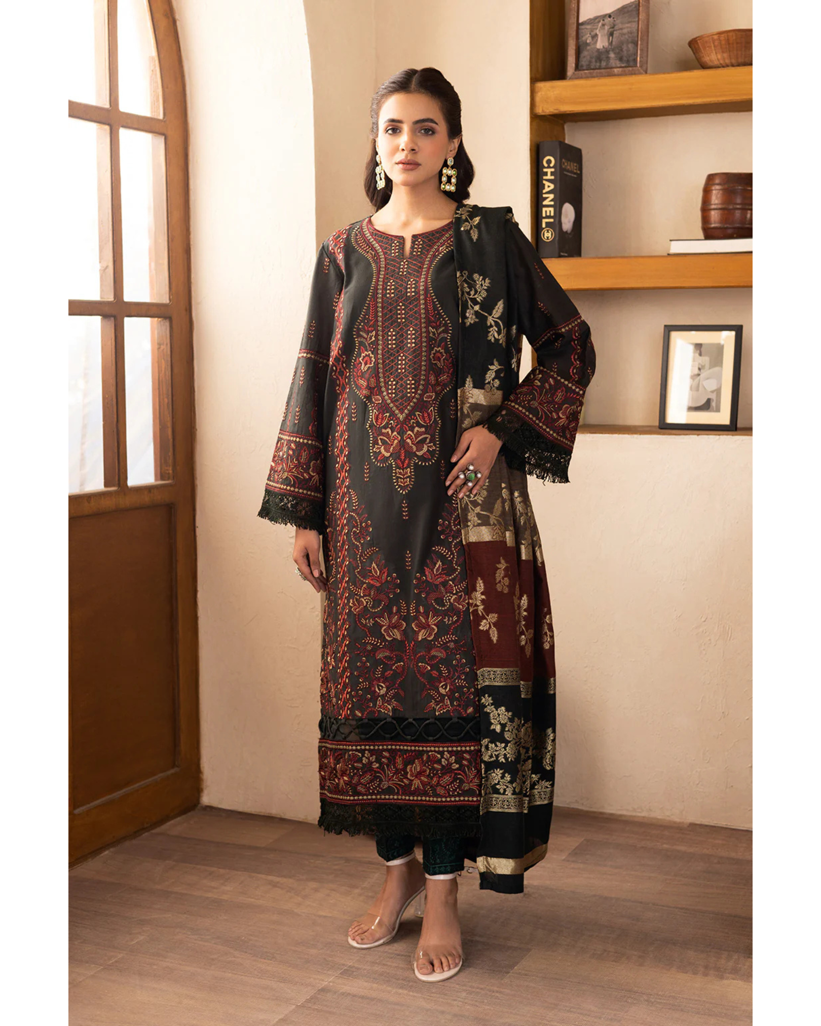 Woman wearing a traditional outfit with intricate patterns in a room with wooden shelves.