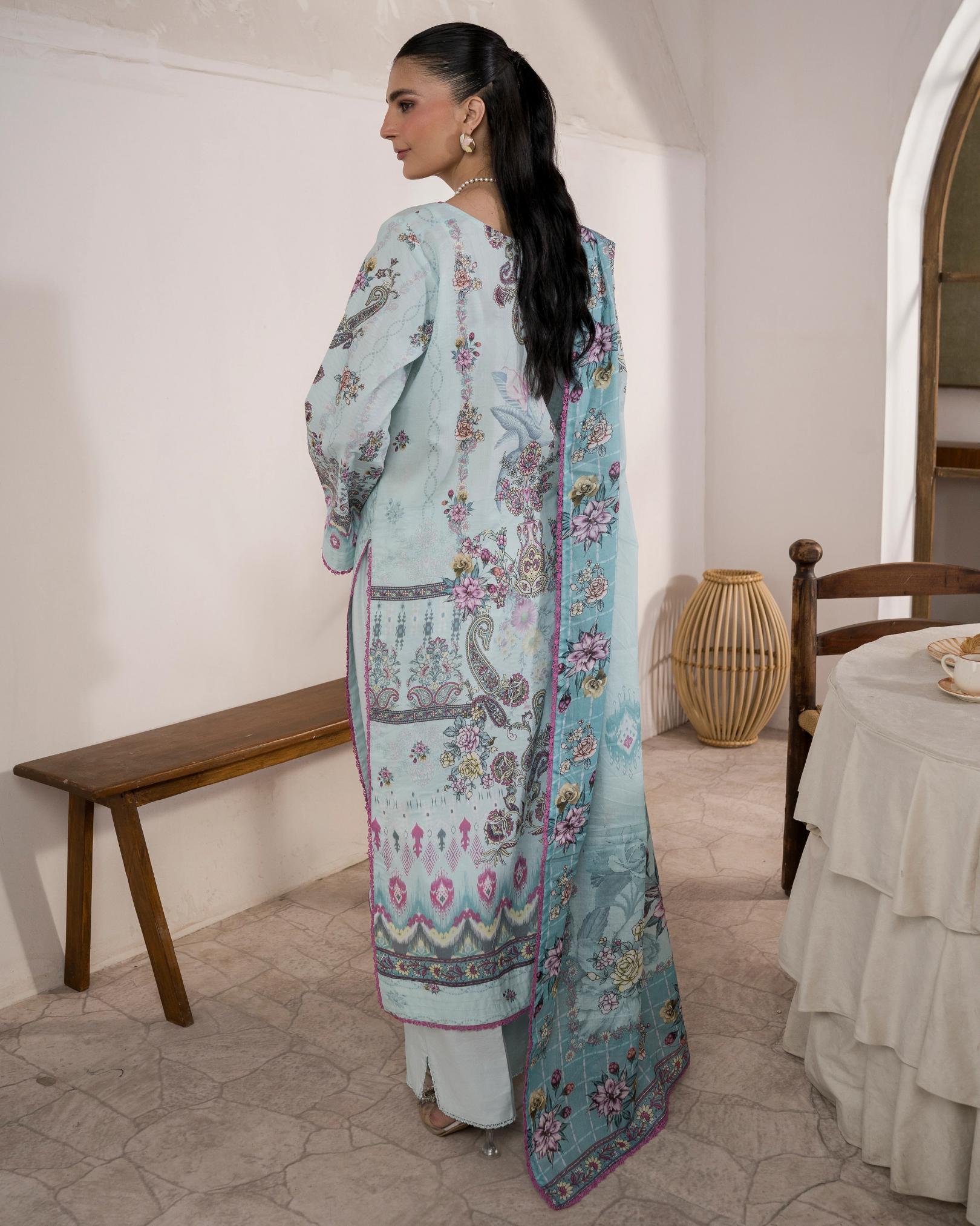 Woman in a light blue floral traditional outfit standing in a room with wooden furniture and a table.