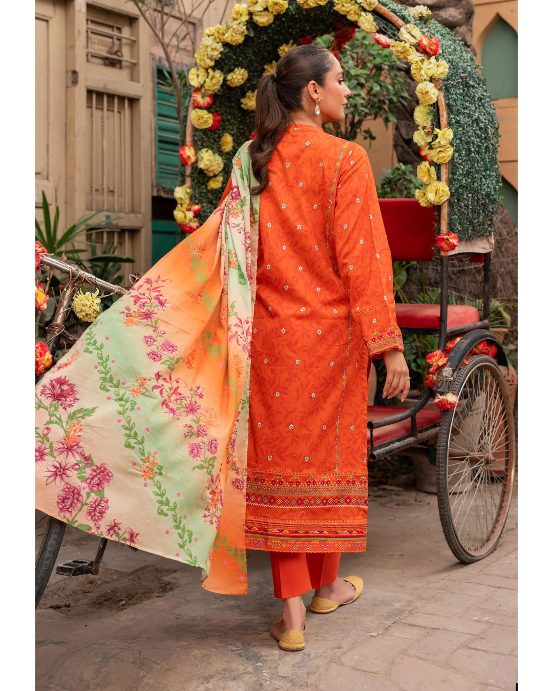 Woman in an orange traditional outfit with a floral dupatta standing next to a decorated entrance.