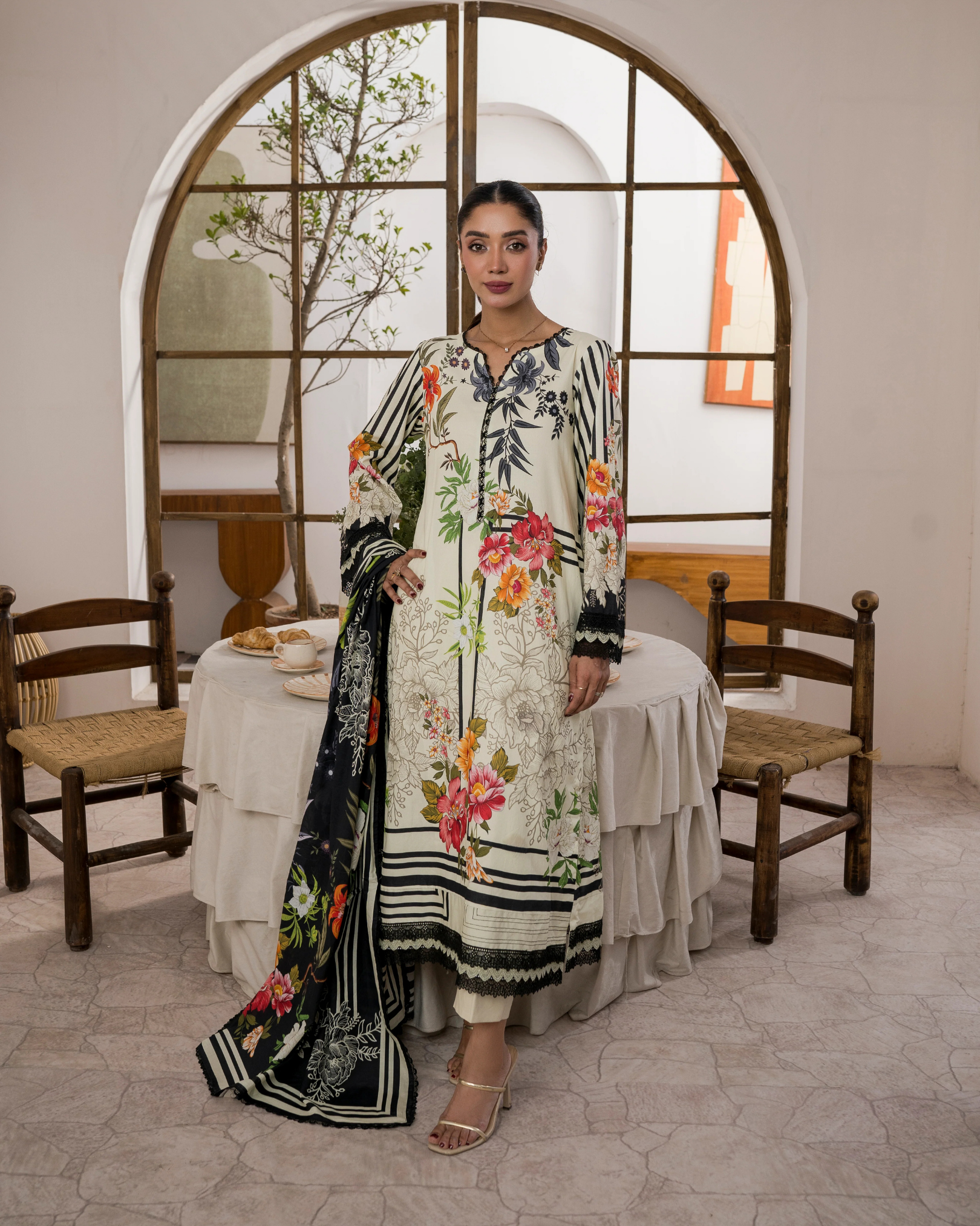 Woman in a floral dress standing in a room with wooden chairs and a table.