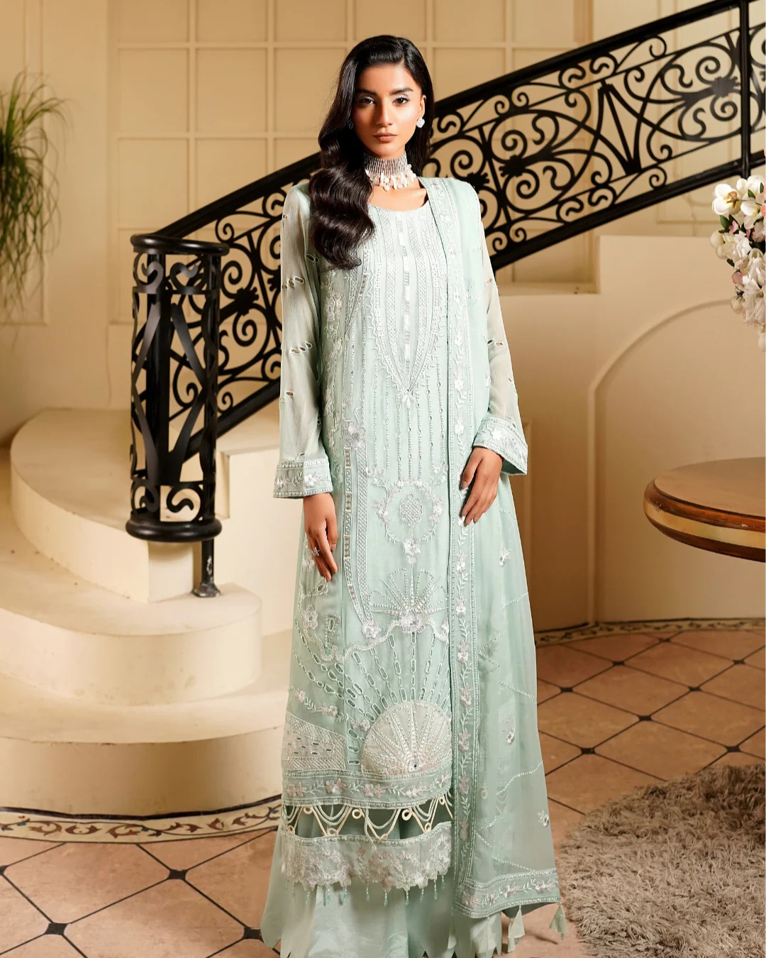 Woman in a light green embroidered outfit standing in a elegant interior setting.