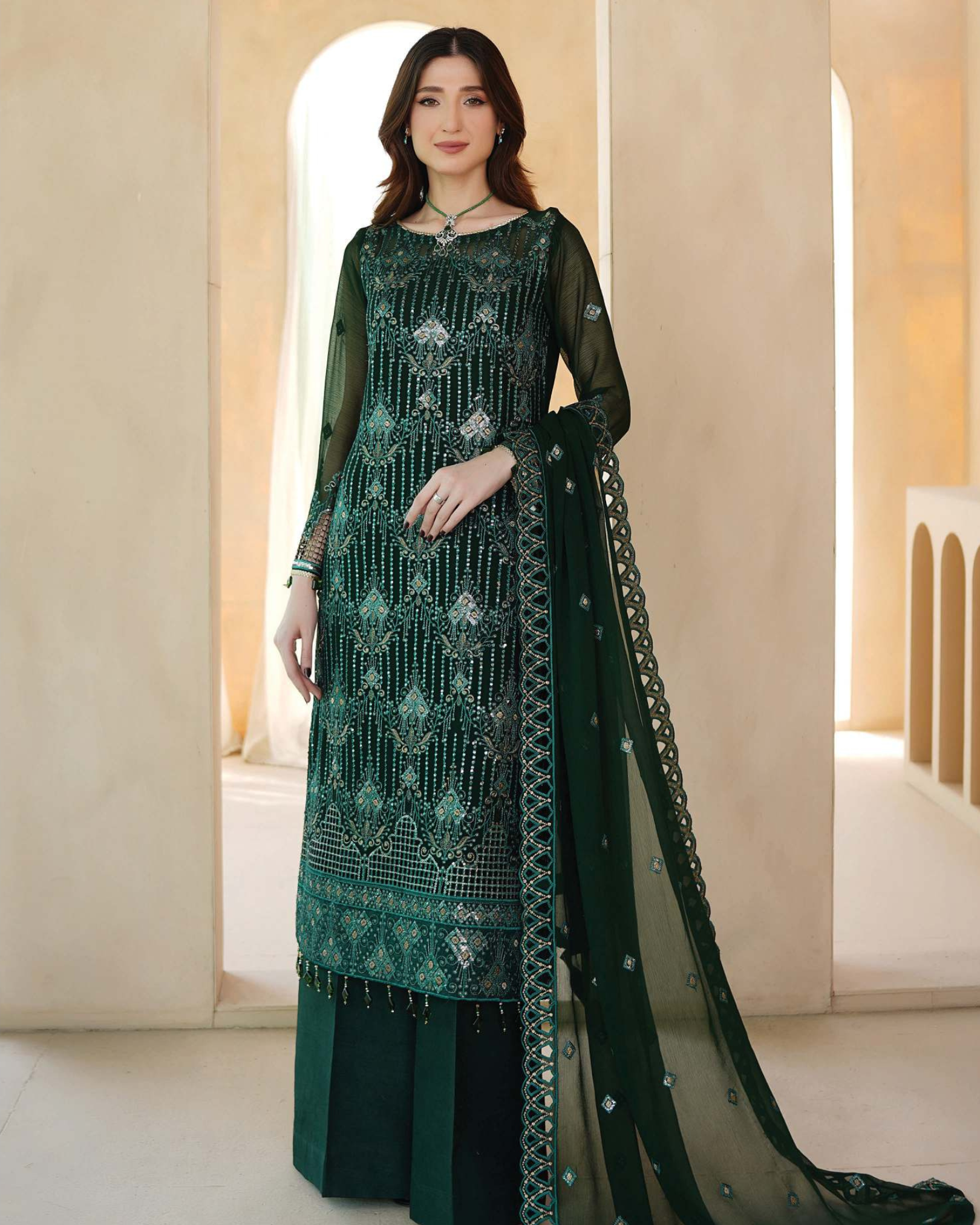 Woman wearing a green embroidered traditional outfit with a sheer dupatta in a neutral setting