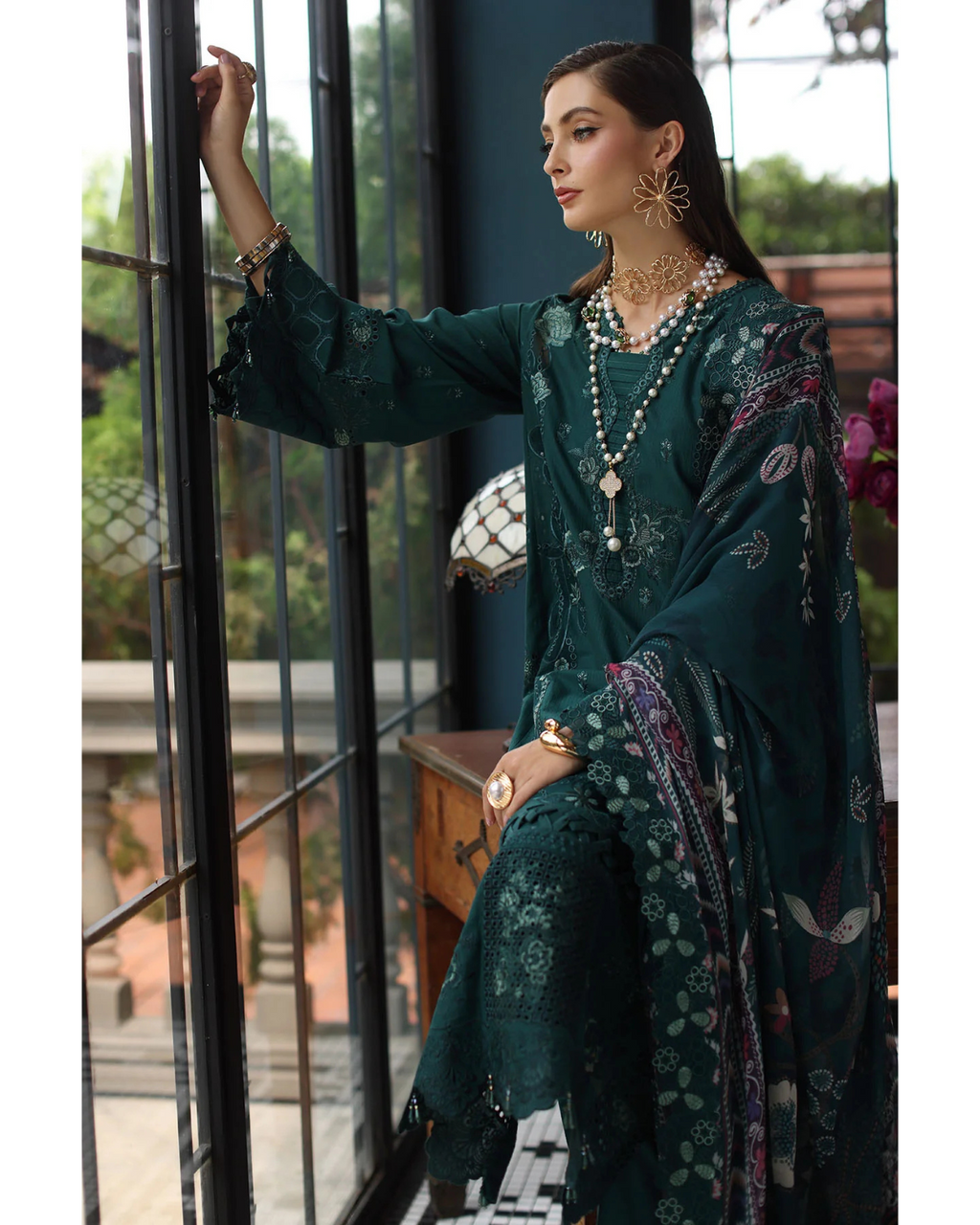 Woman in a dark green traditional outfit with floral patterns, sitting by a window.