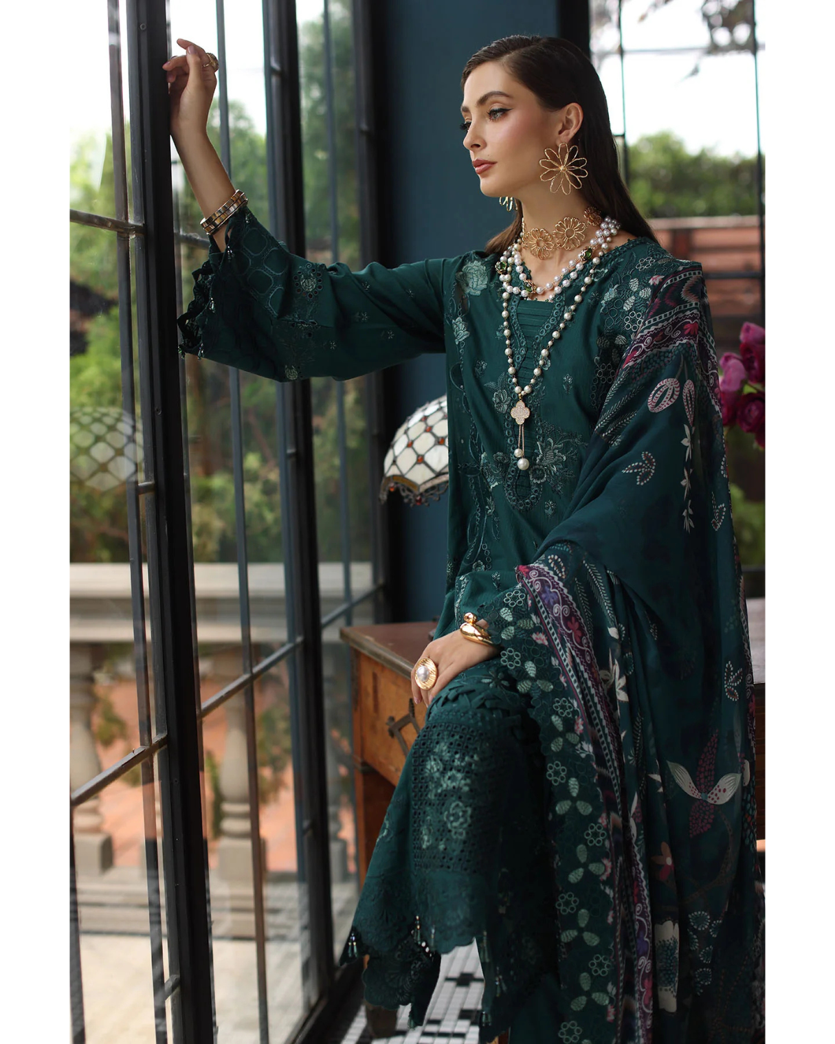 Woman in a dark green traditional outfit with floral patterns, sitting by a window.