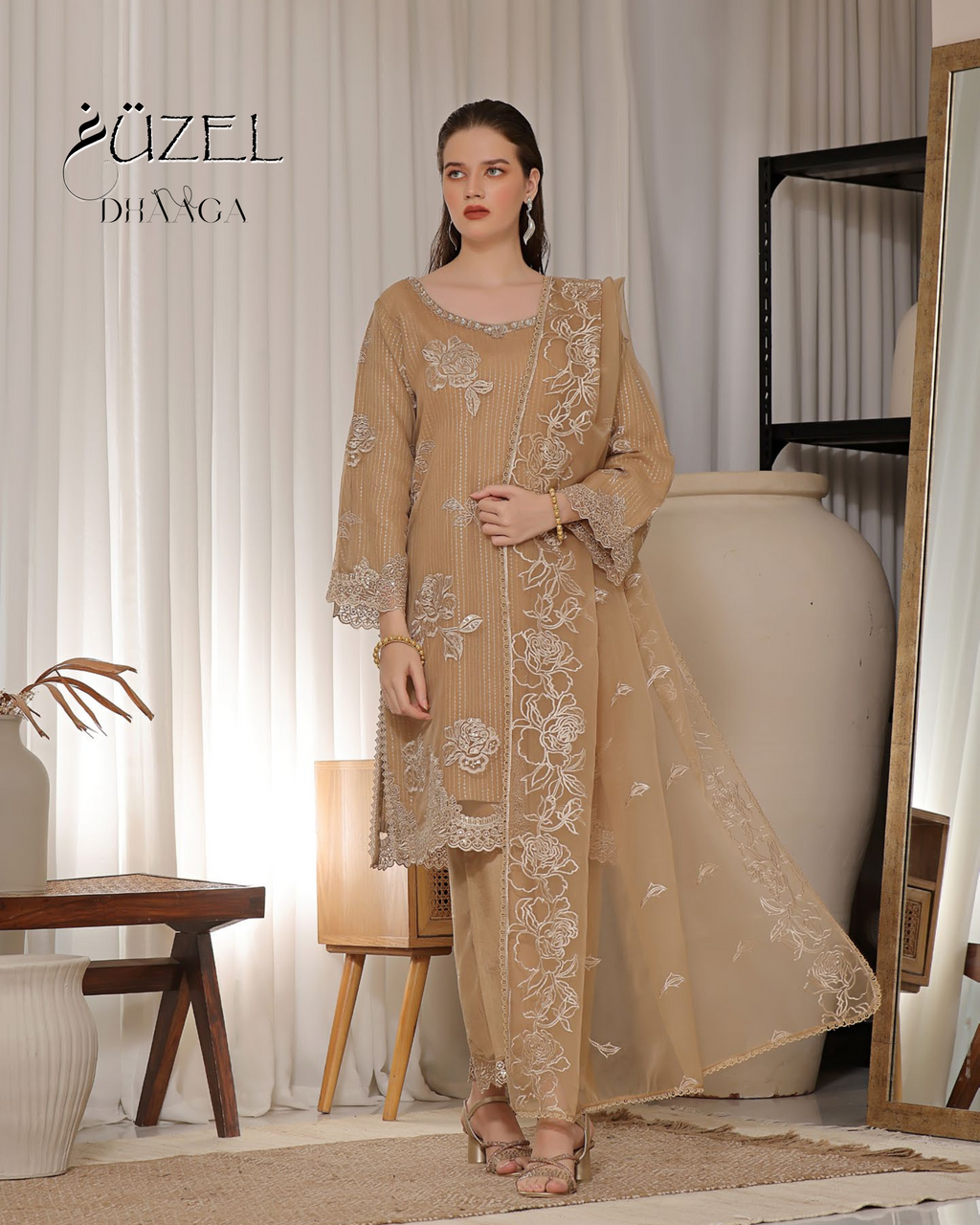 Woman wearing a beige embroidered outfit in a room with white curtains and a wooden table.