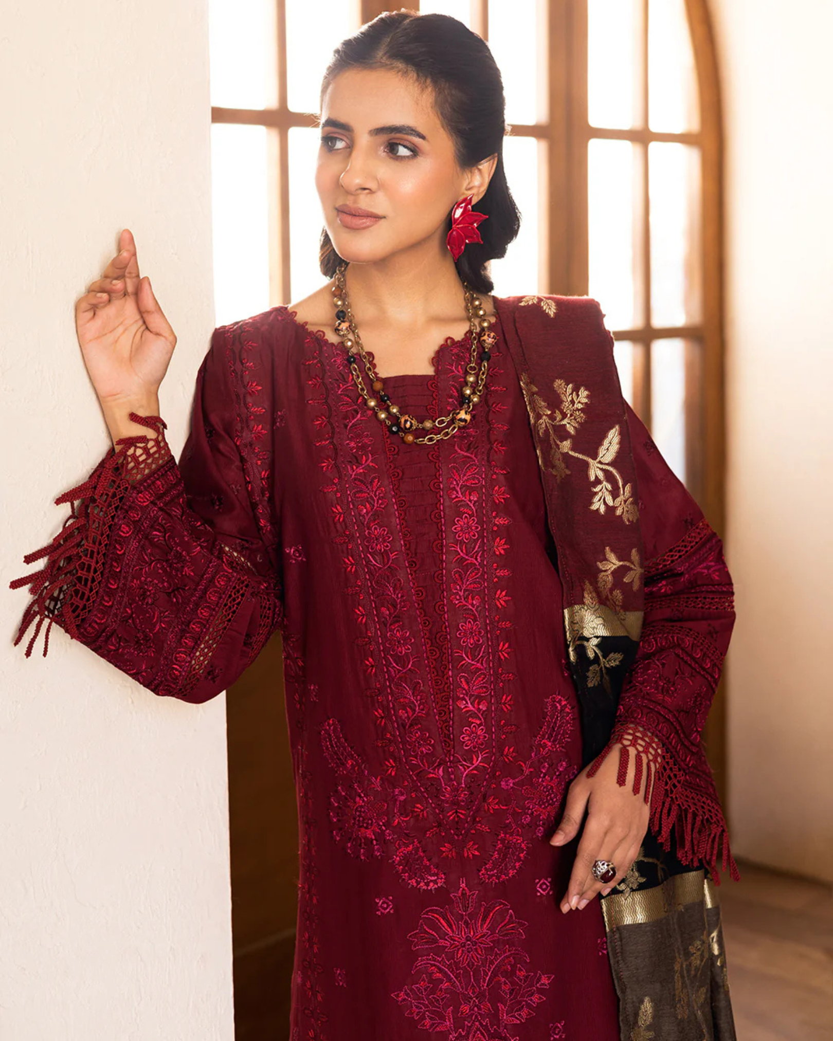 Woman wearing a maroon embroidered outfit with a gold necklace, standing indoors.