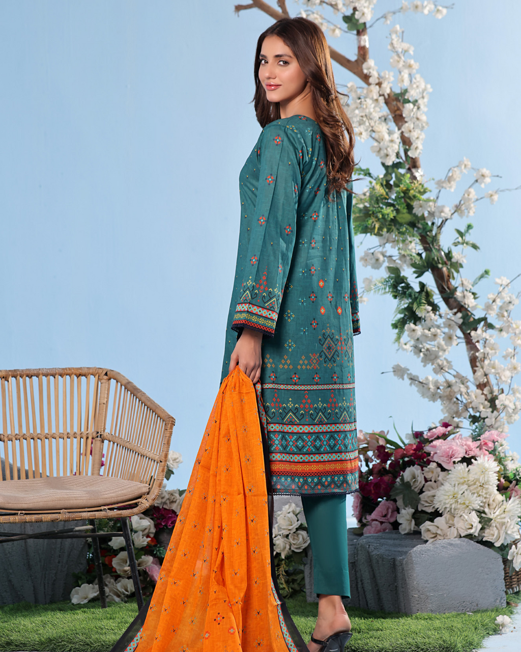Woman in teal embroidered dress with orange dupatta standing outdoors with flowers and a chair in the background
