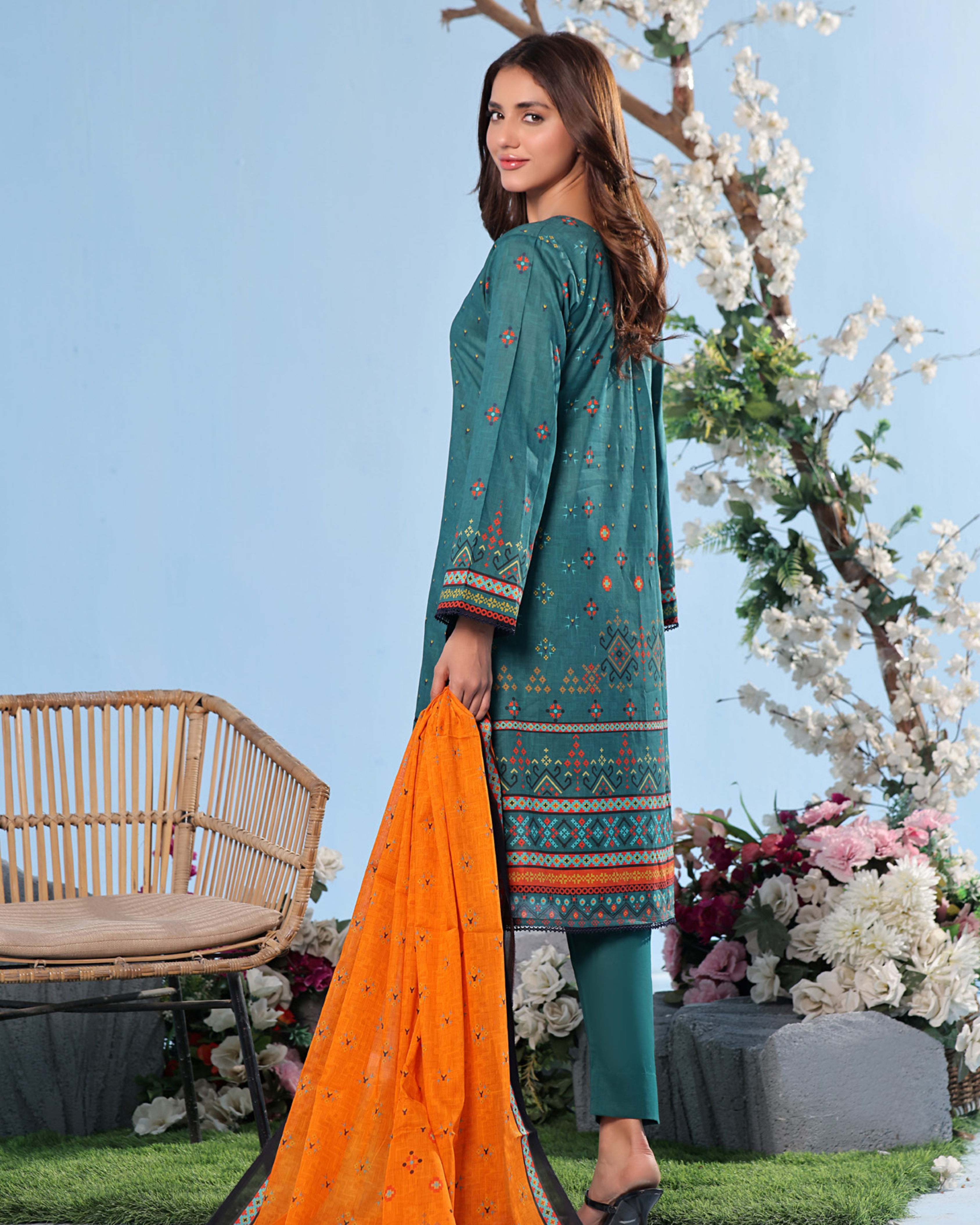 Woman in teal embroidered dress with orange dupatta standing outdoors with flowers and a chair in the background
