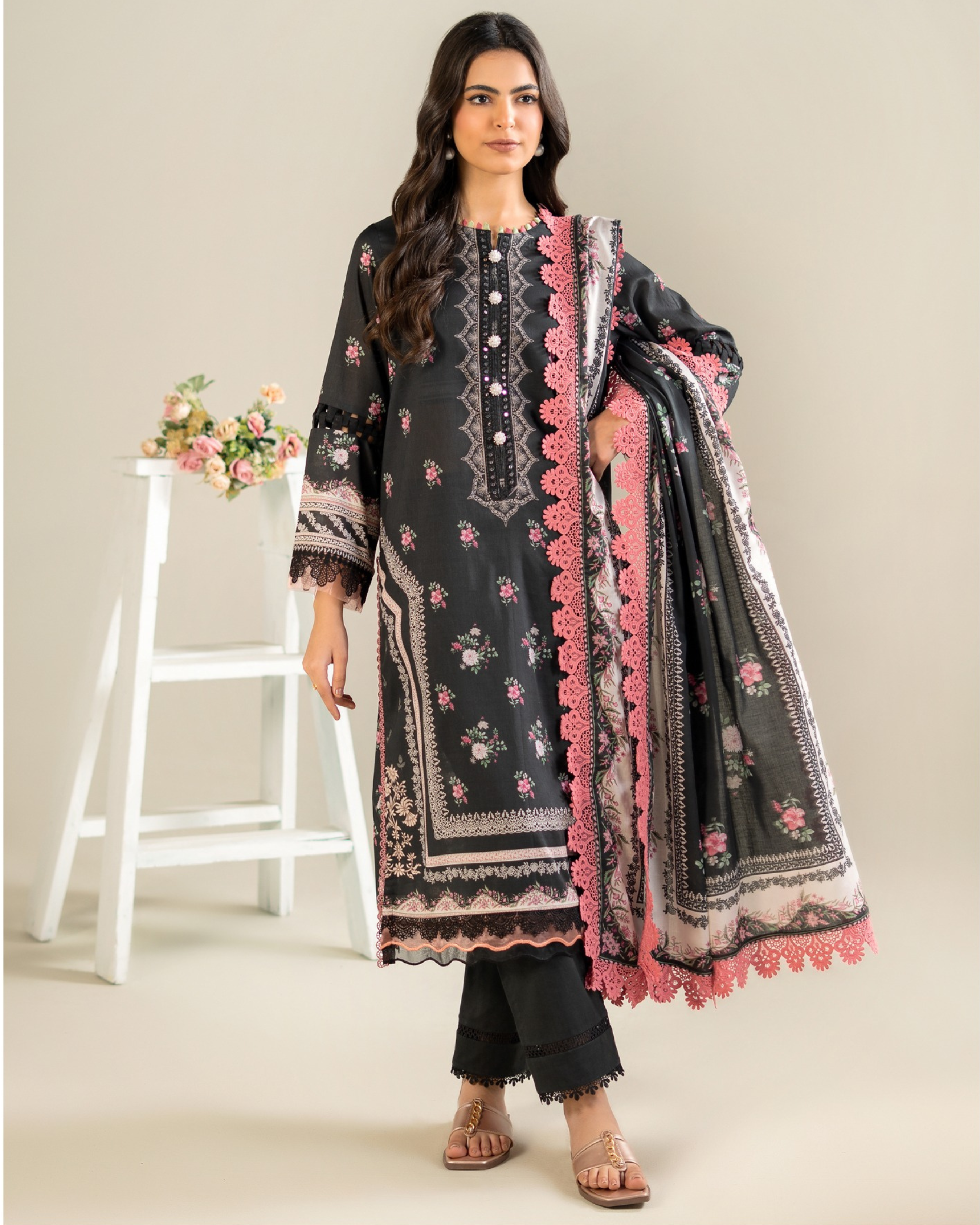 Woman wearing a black and pink embroidered traditional outfit with a white stool and flowers in the background.