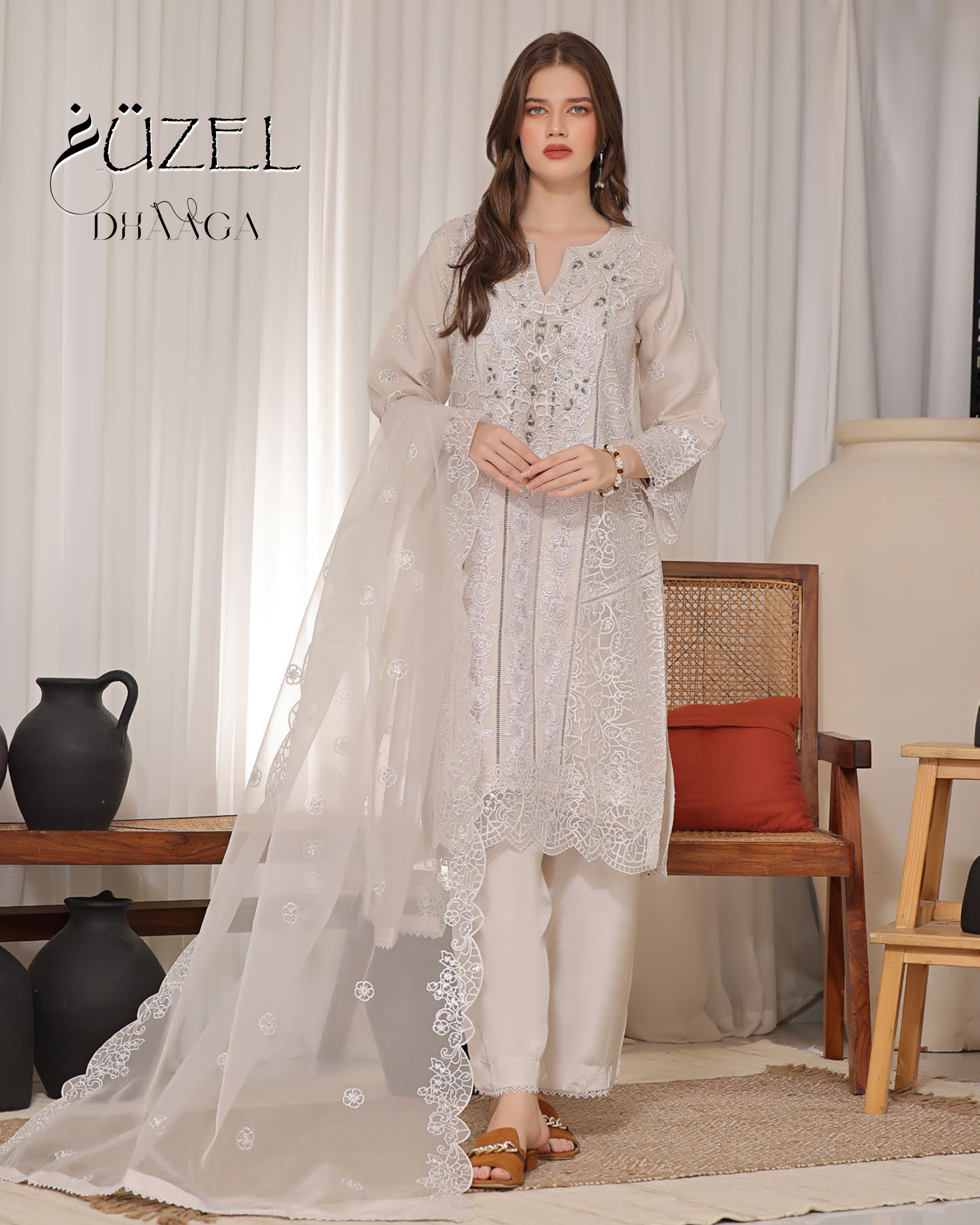 Woman wearing a white embroidered outfit with a chair and decorative items in the background, featuring the brand 'Uzel Dhvaa'.