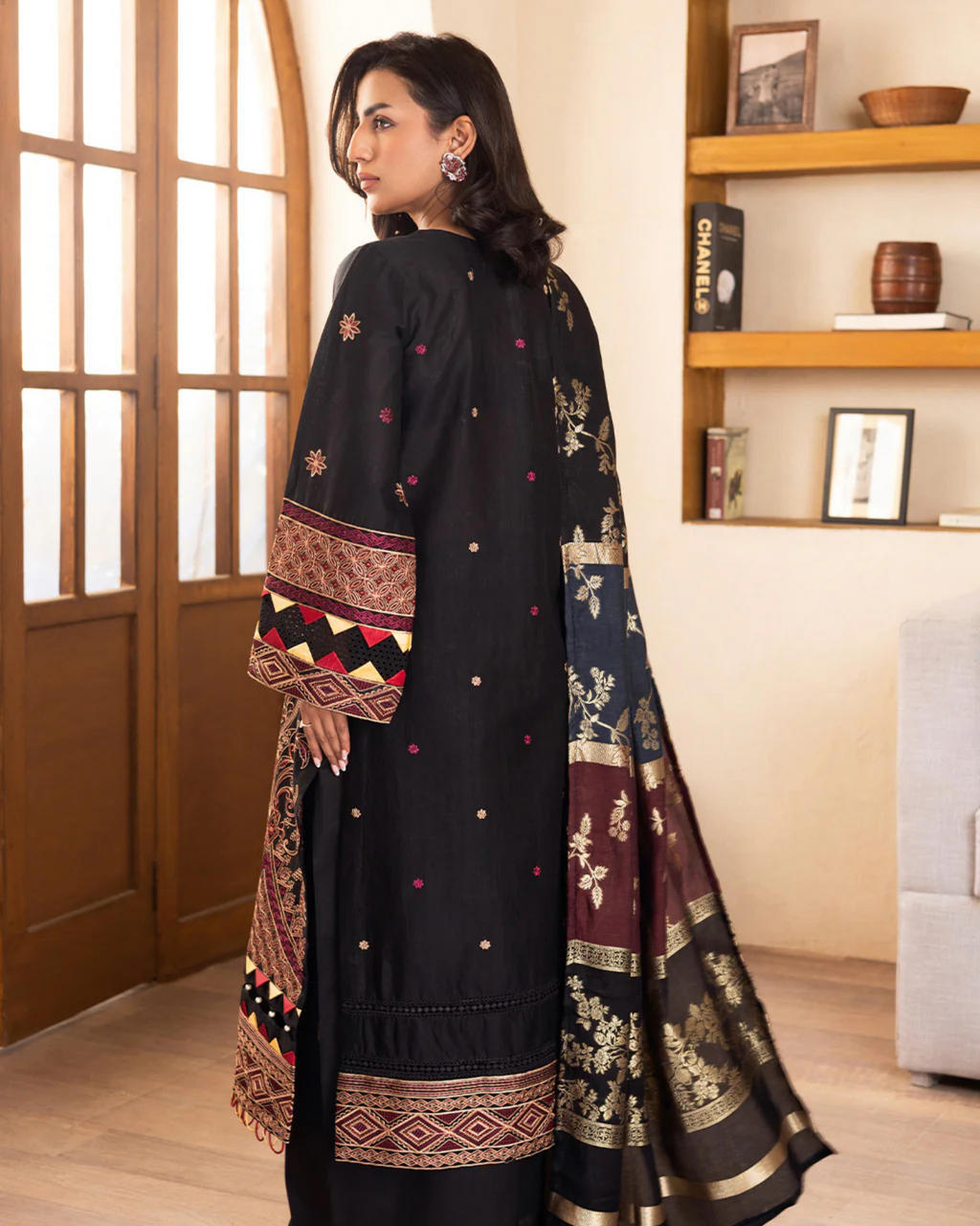 Woman wearing a black traditional outfit with intricate patterns in a room with wooden shelves.