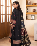 Woman wearing a black traditional outfit with intricate patterns in a room with wooden shelves.
