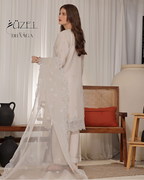 Woman in a light-colored traditional outfit with intricate designs, standing in a room with a wooden chair and decorative items.