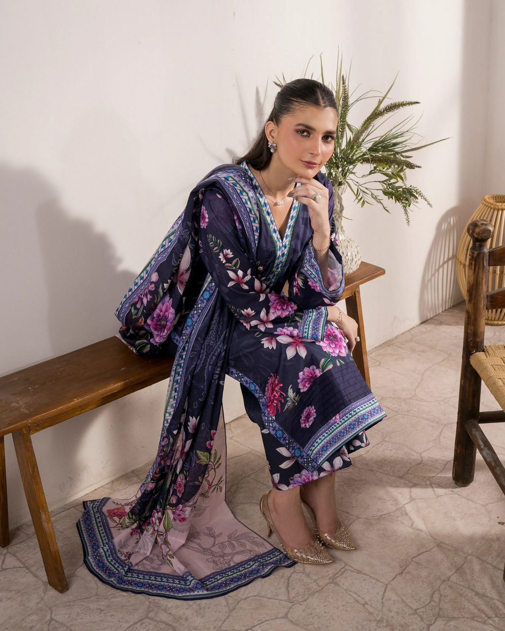 Woman in a floral kimono sitting on a wooden bench with a plant in the background.