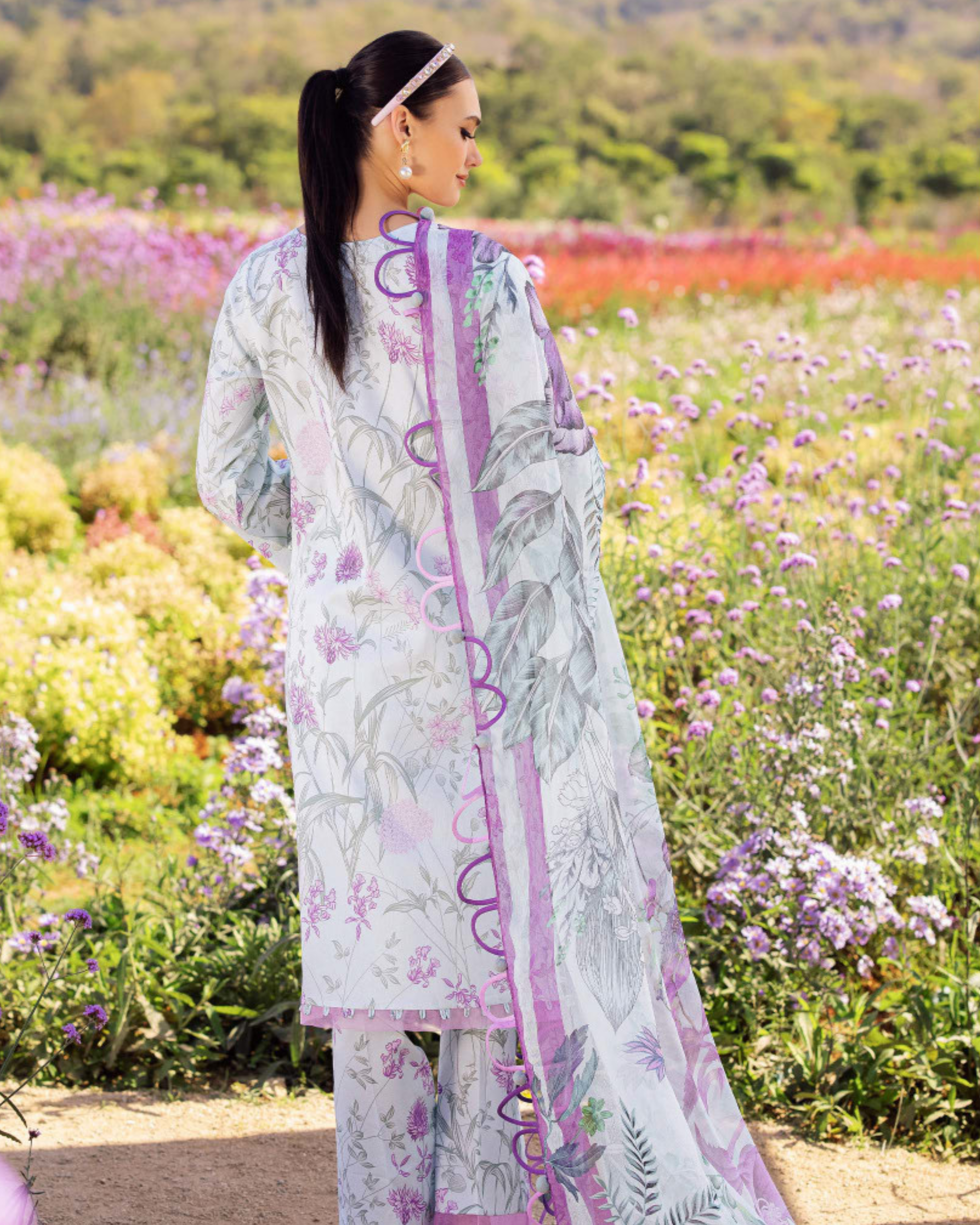 Woman in a light-colored traditional outfit with purple accents standing in a field of flowers.