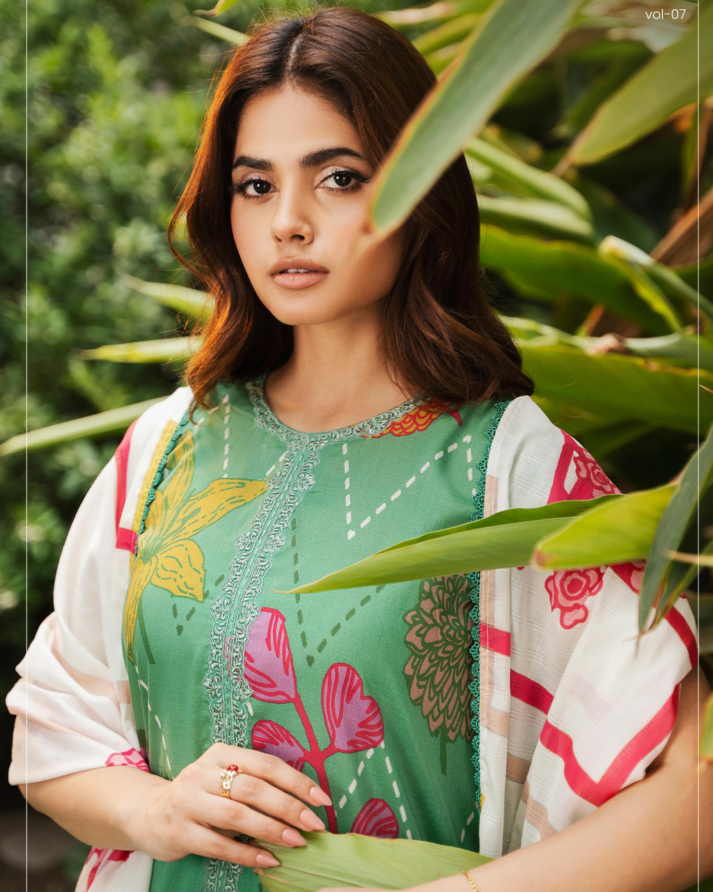 Woman wearing a green embroidered traditional outfit with floral patterns, standing among green leaves.